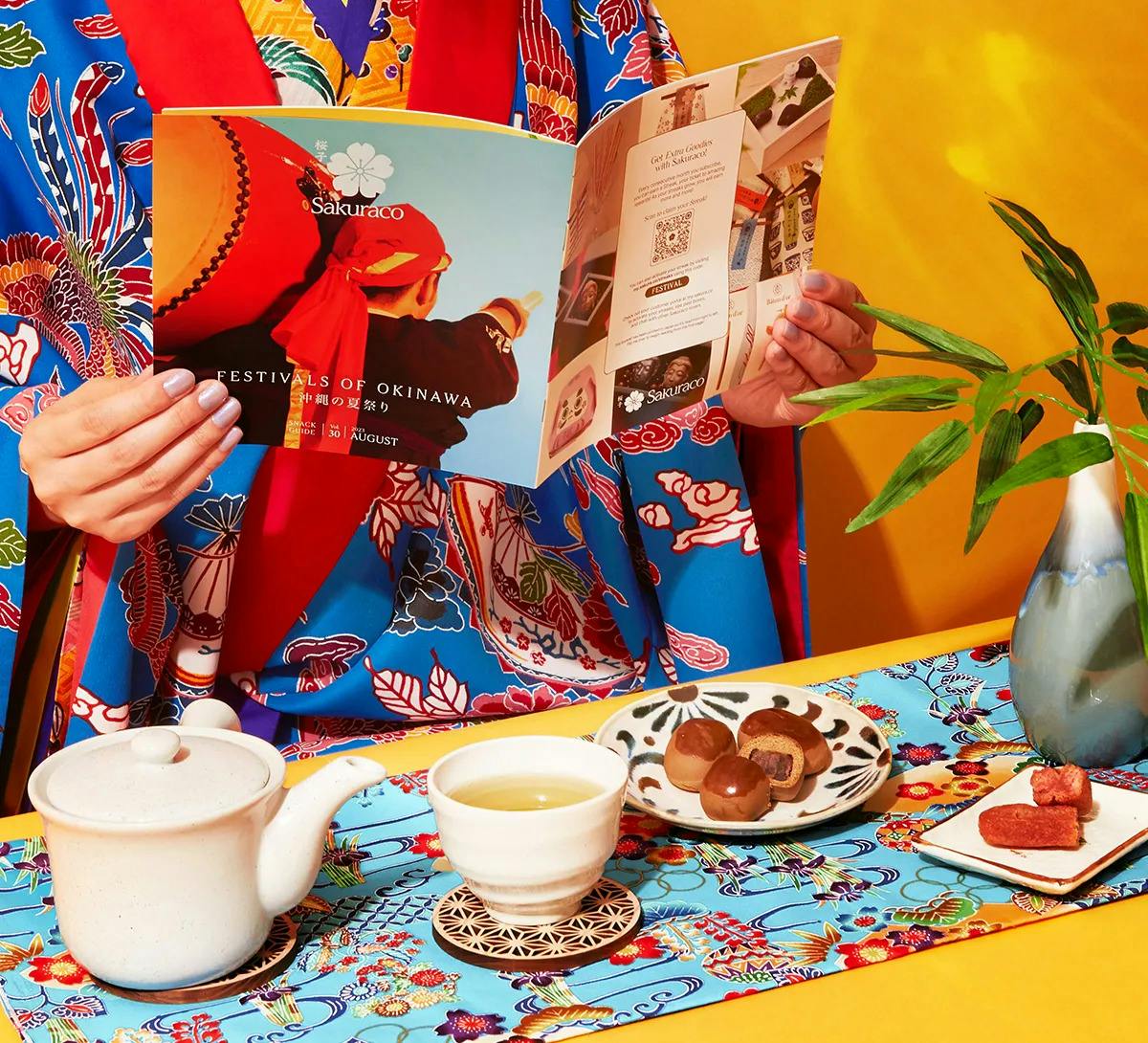 Woman dressed in a vibrant traditional Okinawan Kimono sitting with the August booklet and delectable snacks and tea.