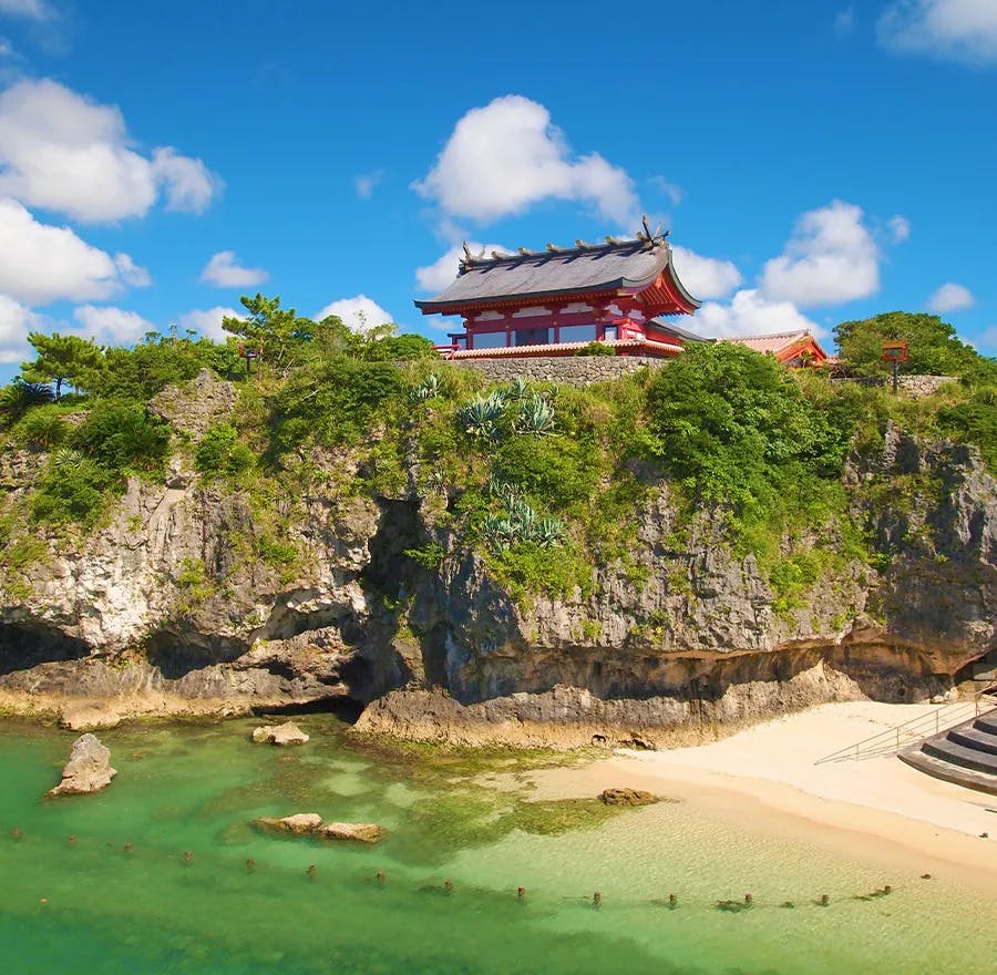An ancient Japanese temple sits on top of a steep cliff surrounded by white sand beaches and clear water.