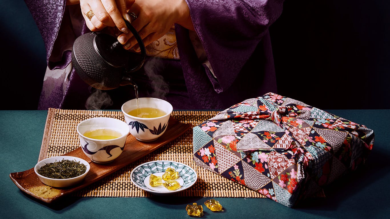 A woman in a purple kimono pours Aged Sencha into a tea cup next to a box wrapped in a furoshiki and a plate of bekko ame.