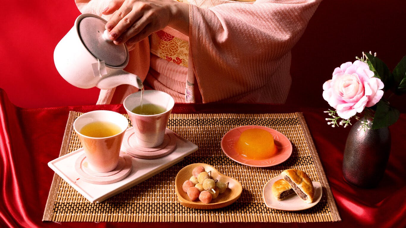 A woman in a pink kimono pours tea surrounded by treats and a rose