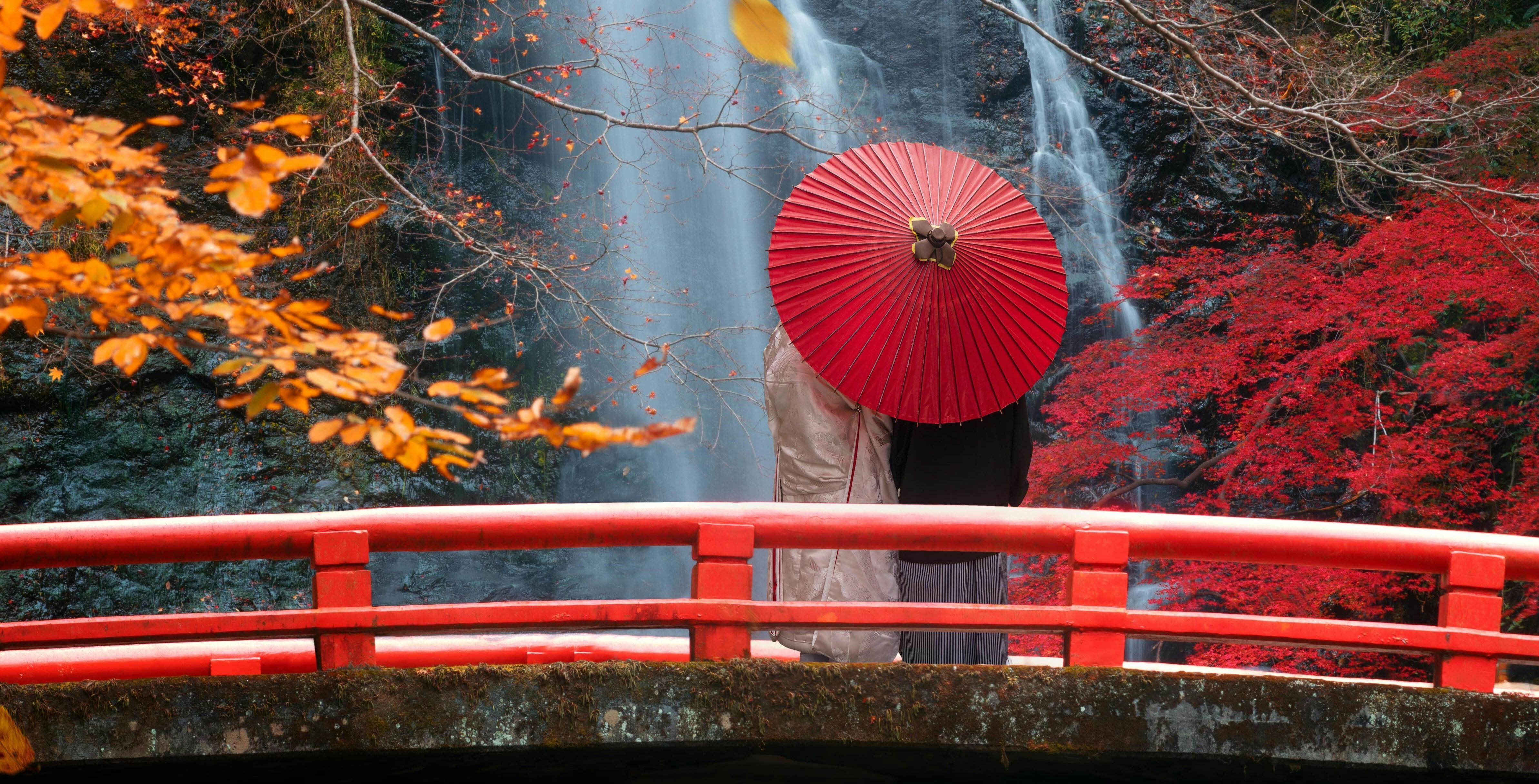 A couple dressed in traditional japanese clothing stand on a red bridge looking at a waterfall