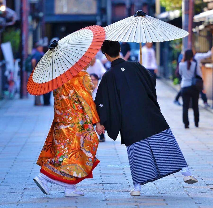 A couple dress in traditional kimonos walk down the street together, holding hands & umbrellas.