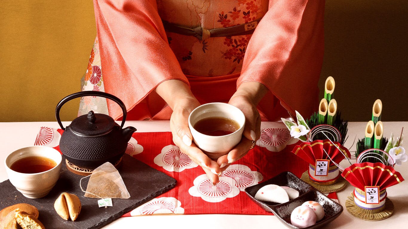 A woman in a pink kimono holds Genmai Black Bean Houjicha surrounded by New Years decorations and assorted Japanese traditional food.