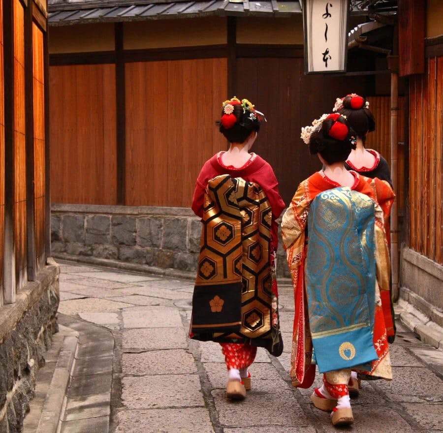 A photo of two geisha walking through the streets of Gion, surrounded by old wooden machiya tea houses