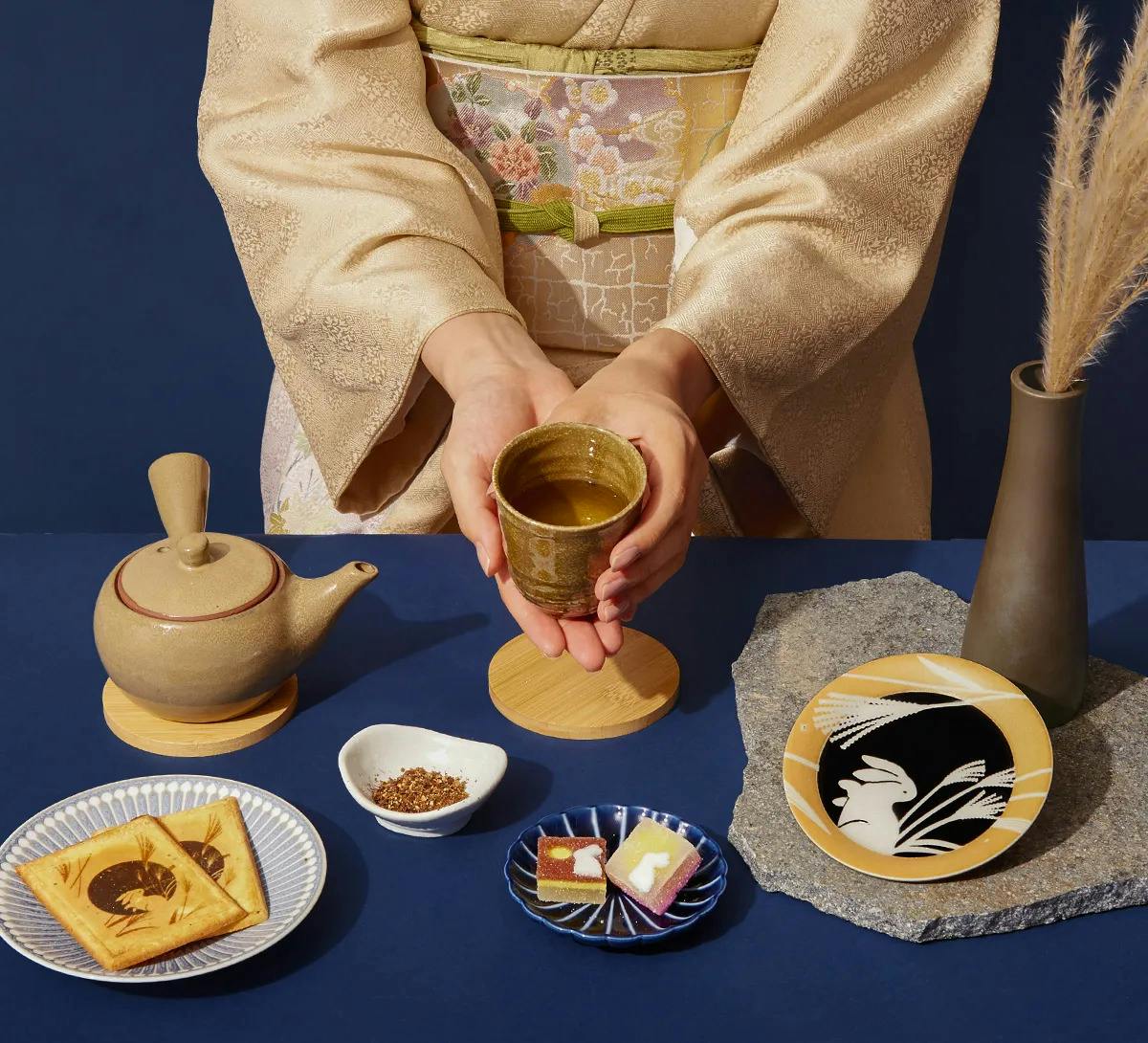 A woman dressed in a kimono sits pouring tea, with traditional snacks