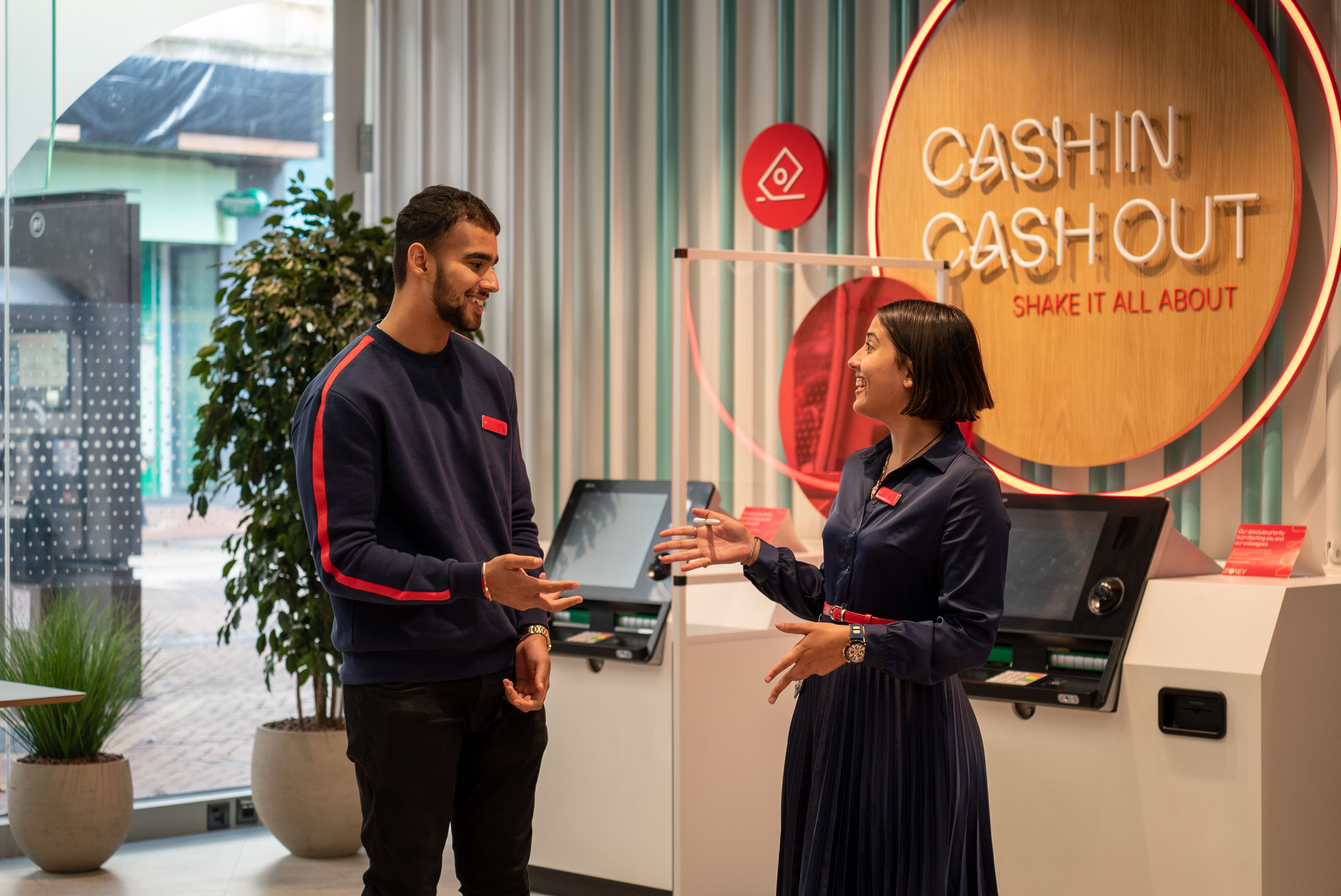 two smiling Virgin Money store employees. They are chatting by the cash machines in store.