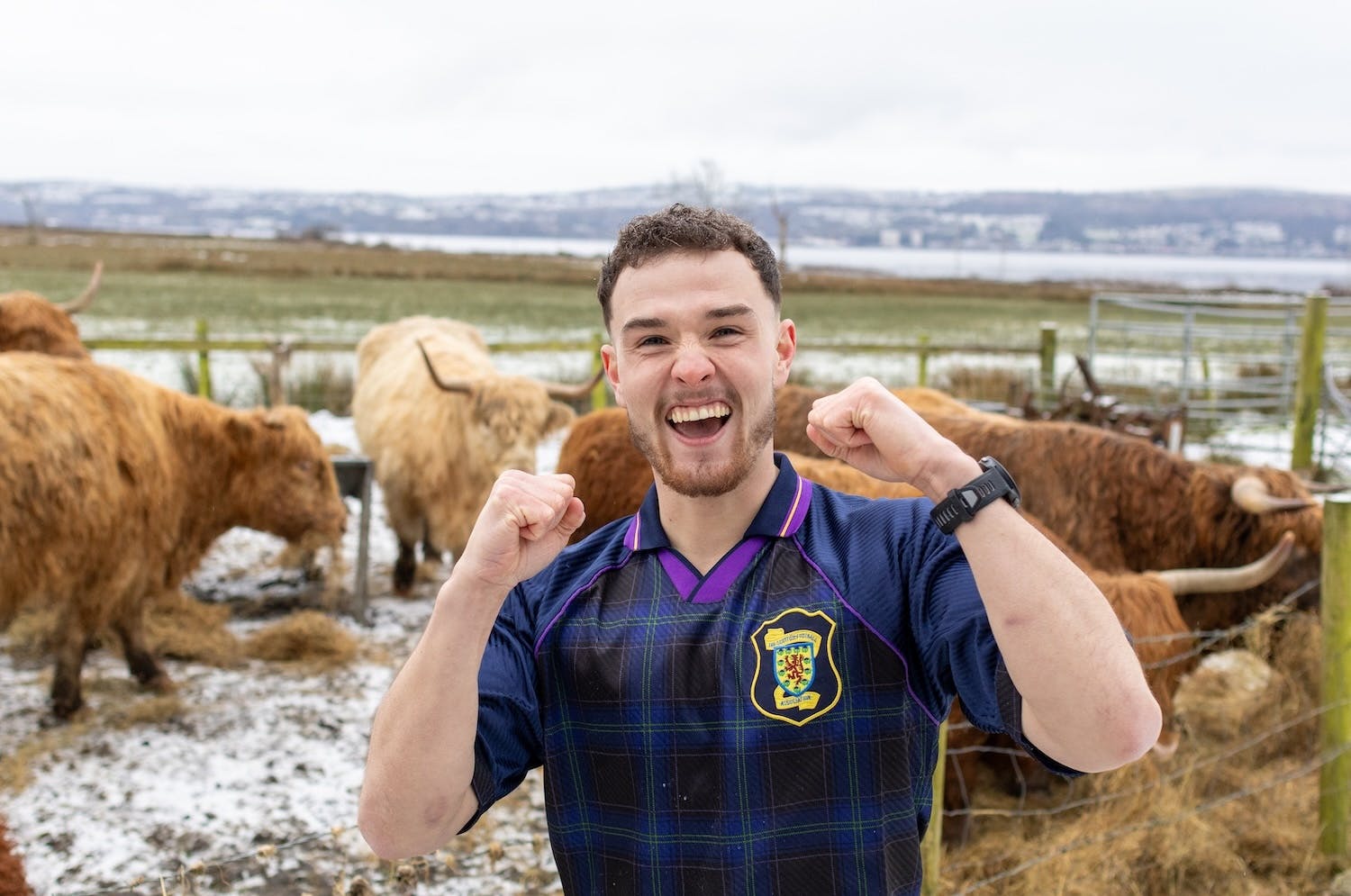 Craig standing in a snowy field posing with raised fists, surrounded by Highland cows.