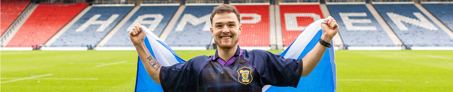 Craig stands on a football pitch holding a large Scottish flag outstretched behind them. He is wearing a dark sports jersey with a crest on the chest, and the stadium seats in the background spell out “HAMPDEN” in bold letters.