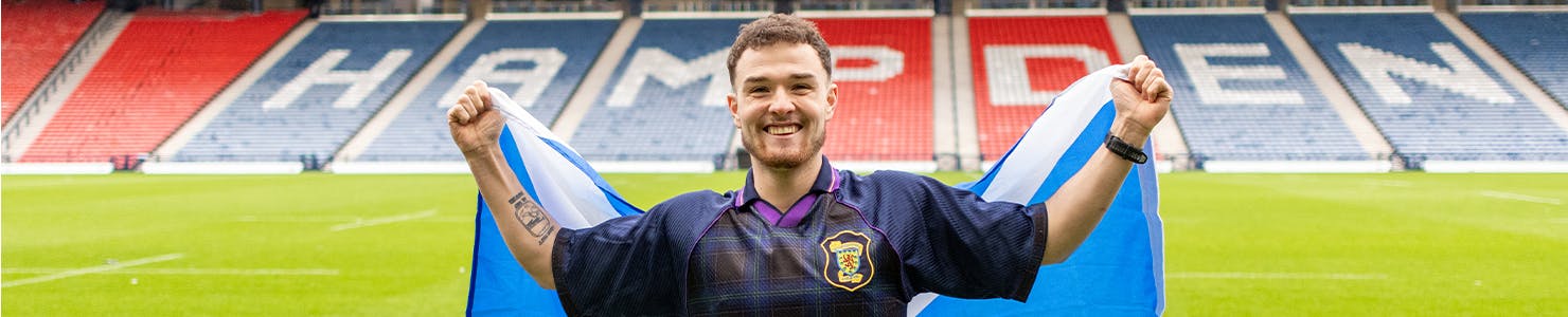 Craig stands on a football pitch holding a large Scottish flag outstretched behind them. He is wearing a dark sports jersey with a crest on the chest, and the stadium seats in the background spell out “HAMPDEN” in bold letters.