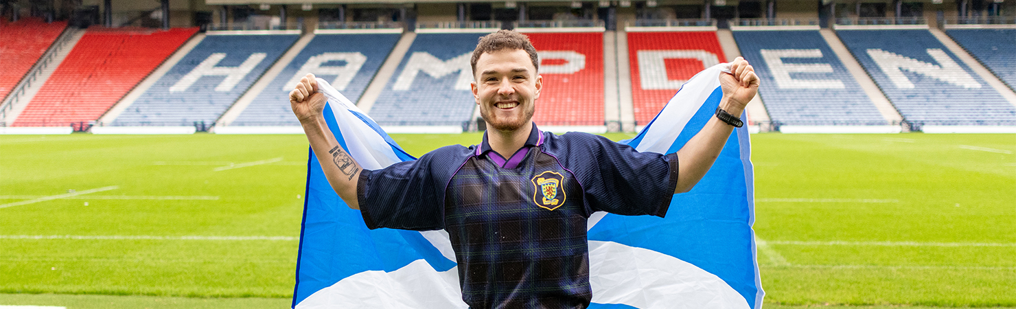 Craig stands on a football pitch holding a large Scottish flag outstretched behind them. He is wearing a dark sports jersey with a crest on the chest, and the stadium seats in the background spell out “HAMPDEN” in bold letters.