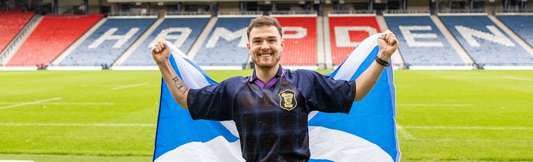 Craig stands on a football pitch holding a large Scottish flag outstretched behind them. He is wearing a dark sports jersey with a crest on the chest, and the stadium seats in the background spell out “HAMPDEN” in bold letters.