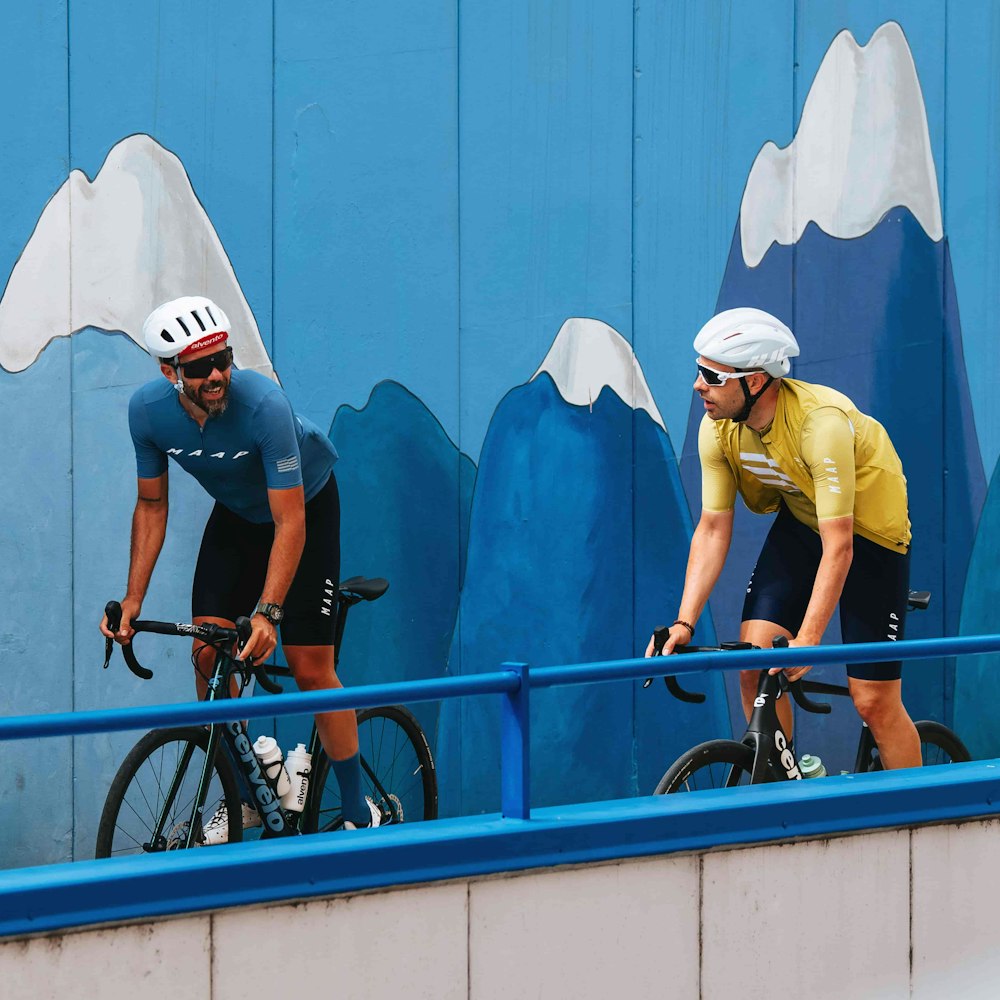 Two cyclists riding with a painted wall of mountains in the background