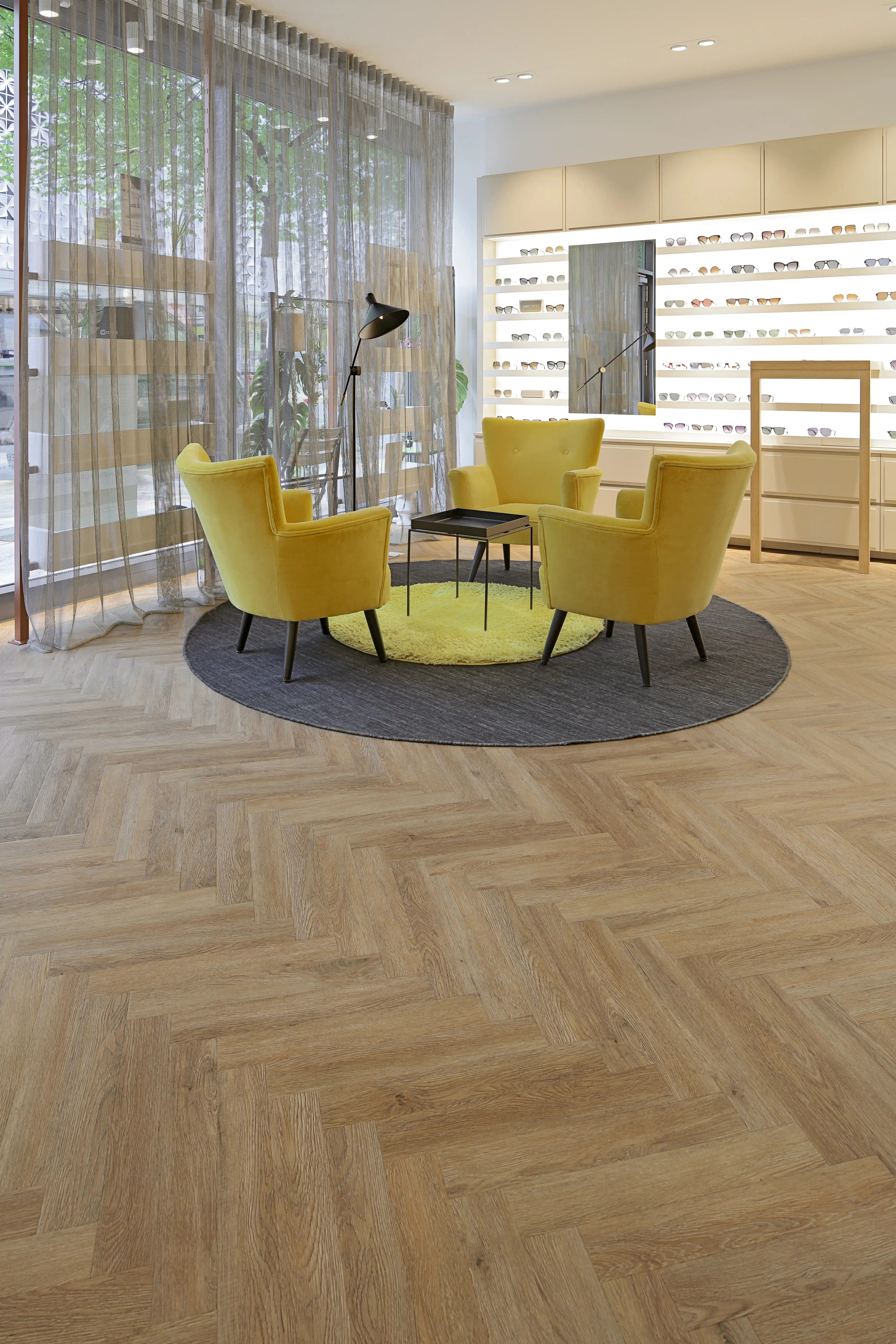 Einladender Loungebereich im Optikergeschäft mit drei gelben Sesseln auf einem runden Teppich, perfekt in Szene gesetzt durch den großflächig verlegten Fischgrät-Vinylboden.