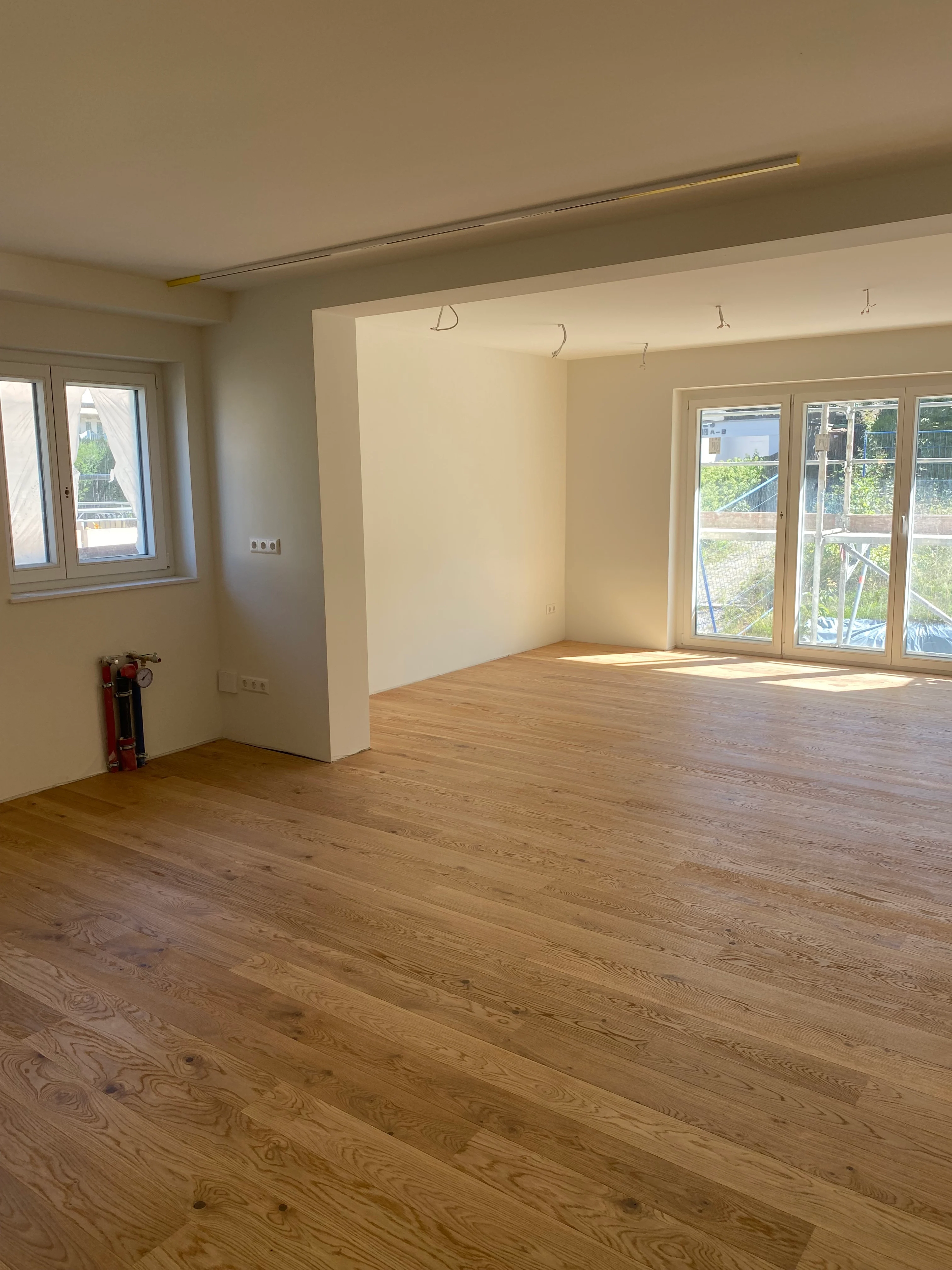 Die natürliche Holzstruktur des weiß geölten Landhausdielen-Parketts wird durch das Sonnenlicht betont, das durch große Fenstertüren fällt. Ein sichtbarer Verteiler an der Wand zeigt den Baufortschritt.