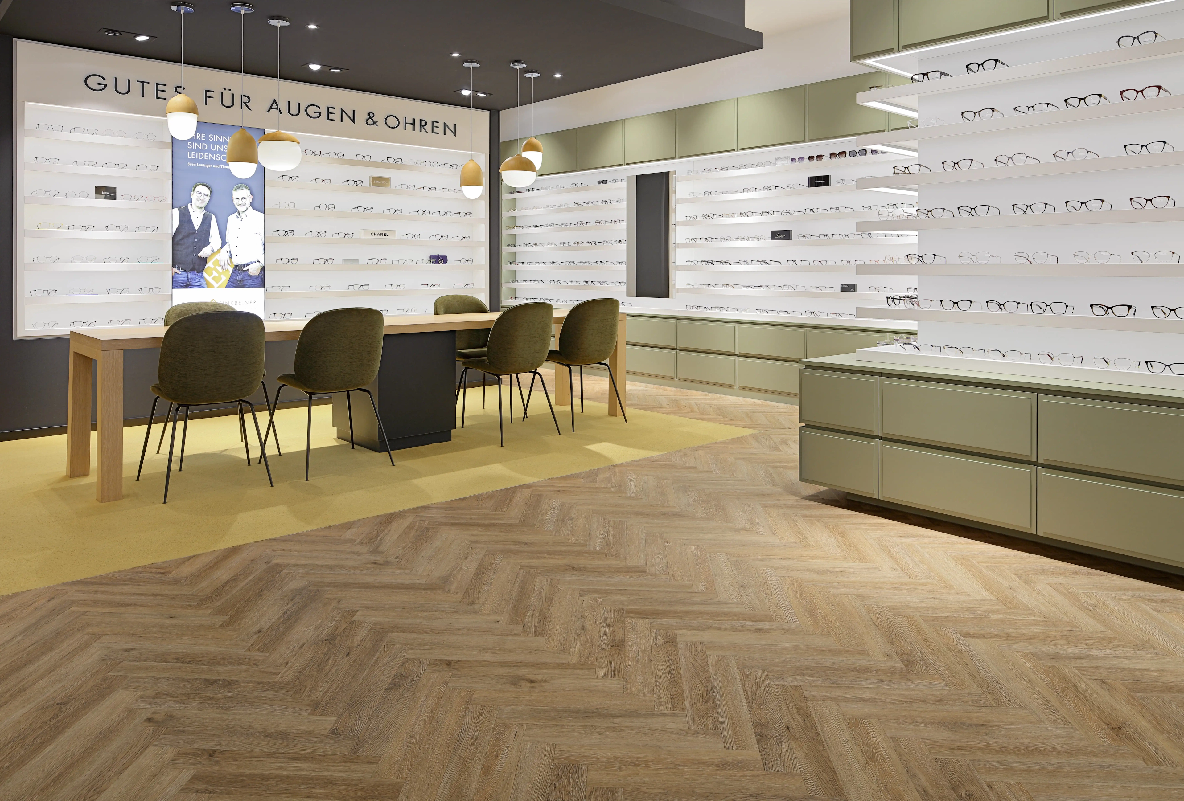 Großzügiger Beratungsbereich im Optikergeschäft in Pforzheim mit einem langen Holztisch auf gelbem Teppichboden, harmonisch ergänzt durch den von Parkett Saussele verlegten Fischgrät-Vinylboden.