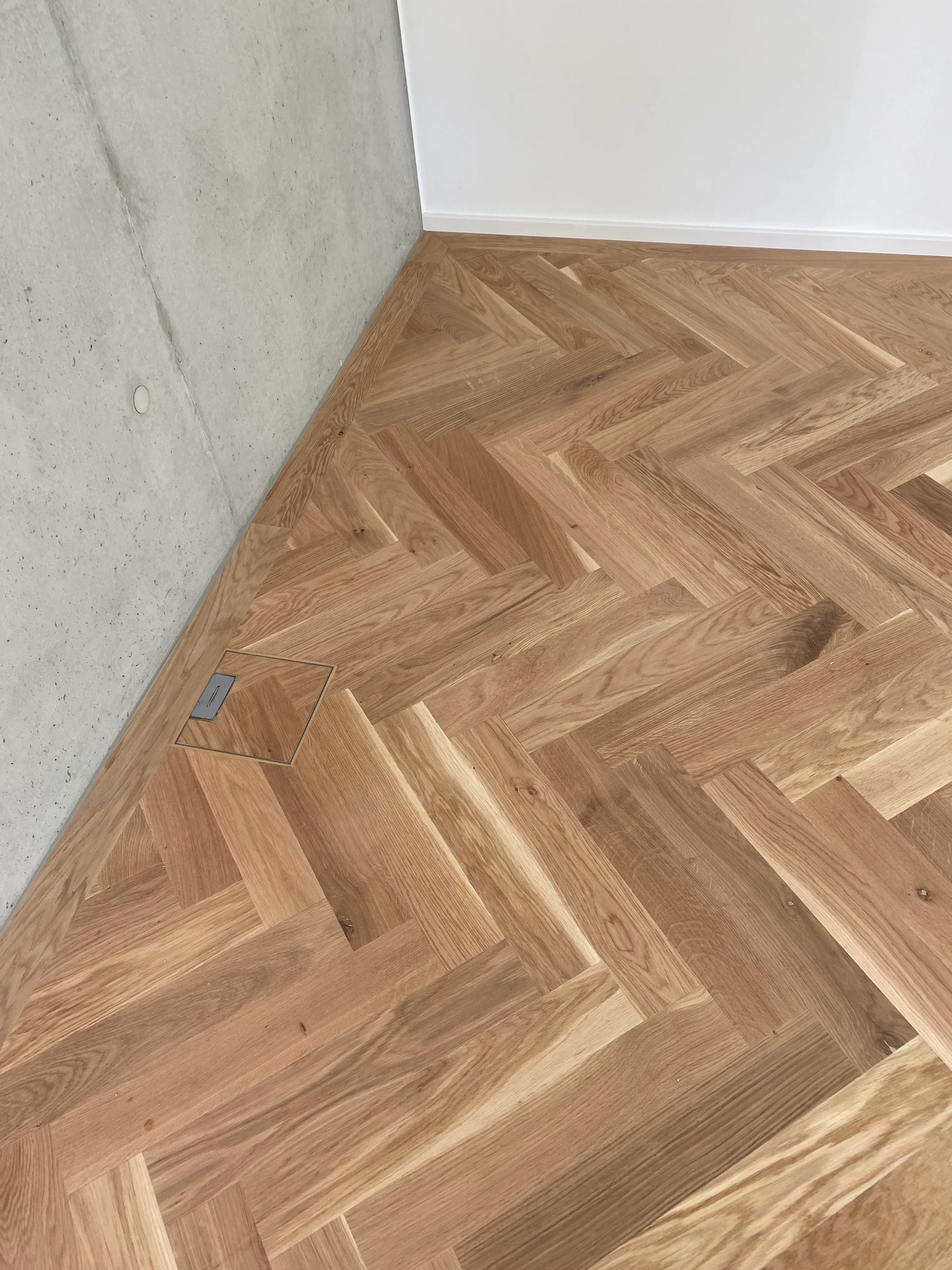 Fischgrätparkett an einer Sichtbetonwand mit einer flächenbündig integrierten Bodensteckdose, handgefertigt von Parkett Saussele.
