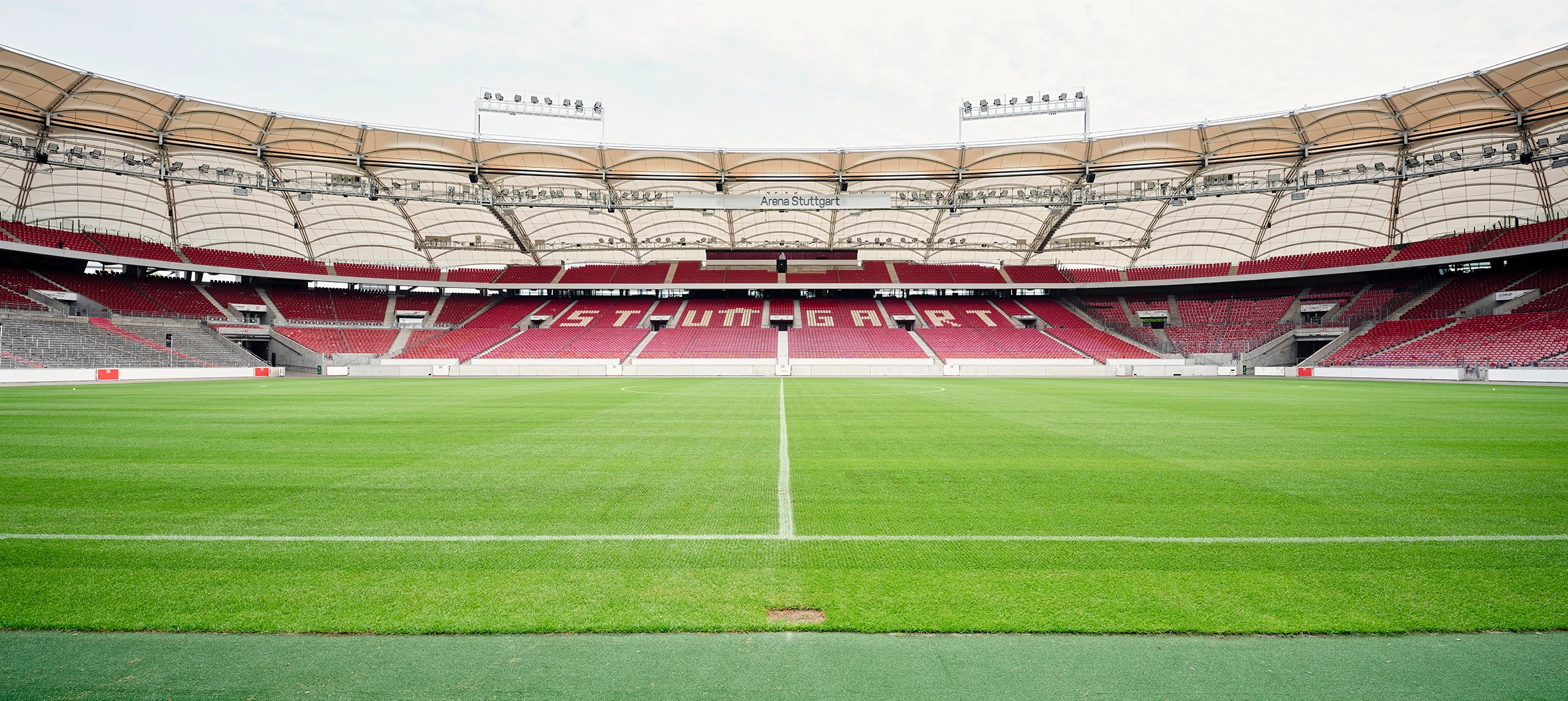 Weites Panorama des grünen Spielfelds und der leeren roten Zuschauerränge in der MHP Arena Stuttgart.