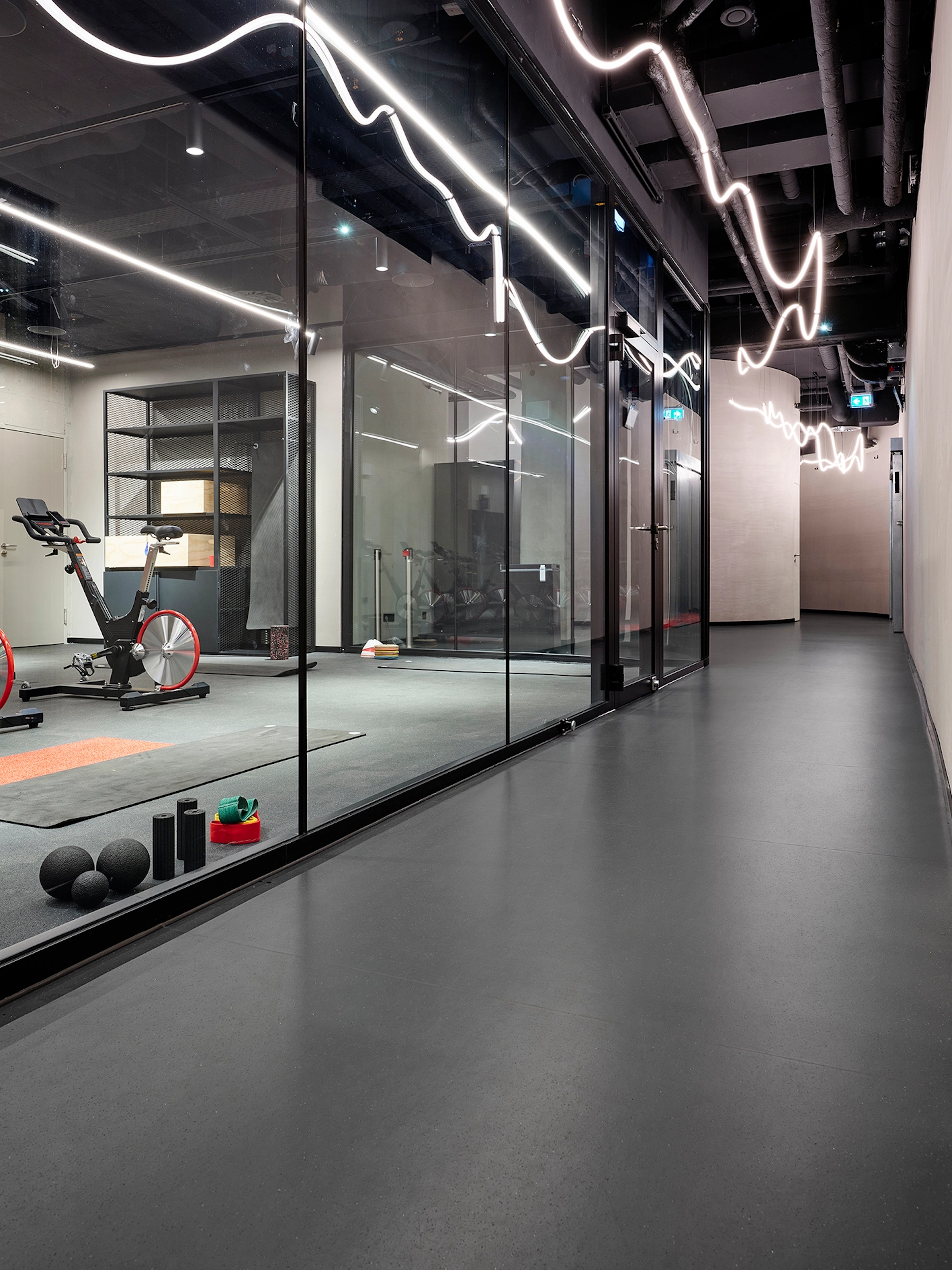 Präzise verlegter dunkelgrauer Boden in einem Flur mit geschwungener Deckenbeleuchtung und Blick durch eine Glaswand in einen Fitnessraum der MHP Arena.