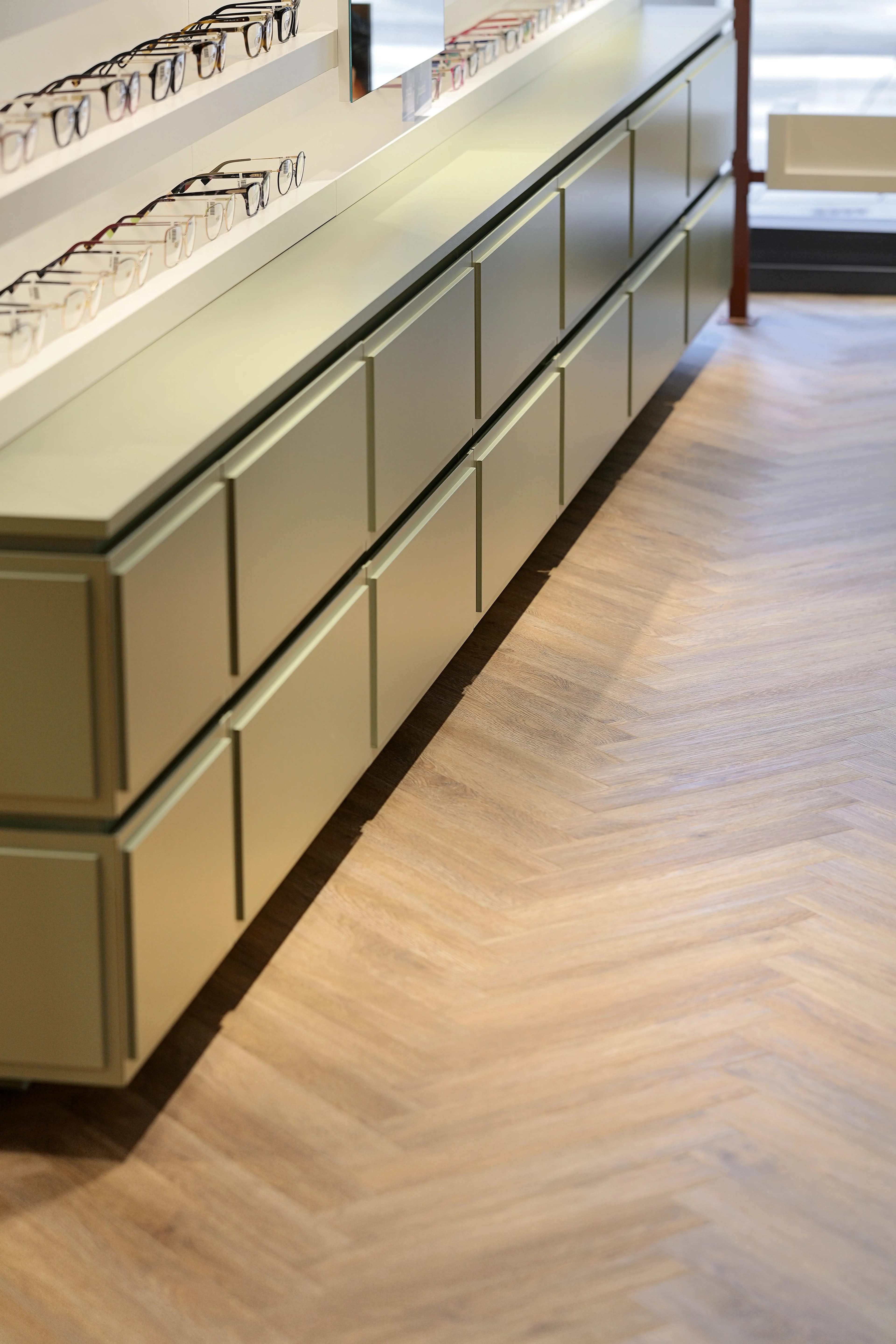 Ein langes, salbeigrünes Präsentationsmöbel mit Brillen im Optikerladen Pforzheim, perfekt kombiniert mit dem von Parkett Saussele verlegten Vinylboden in zeitloser Fischgrät-Optik.