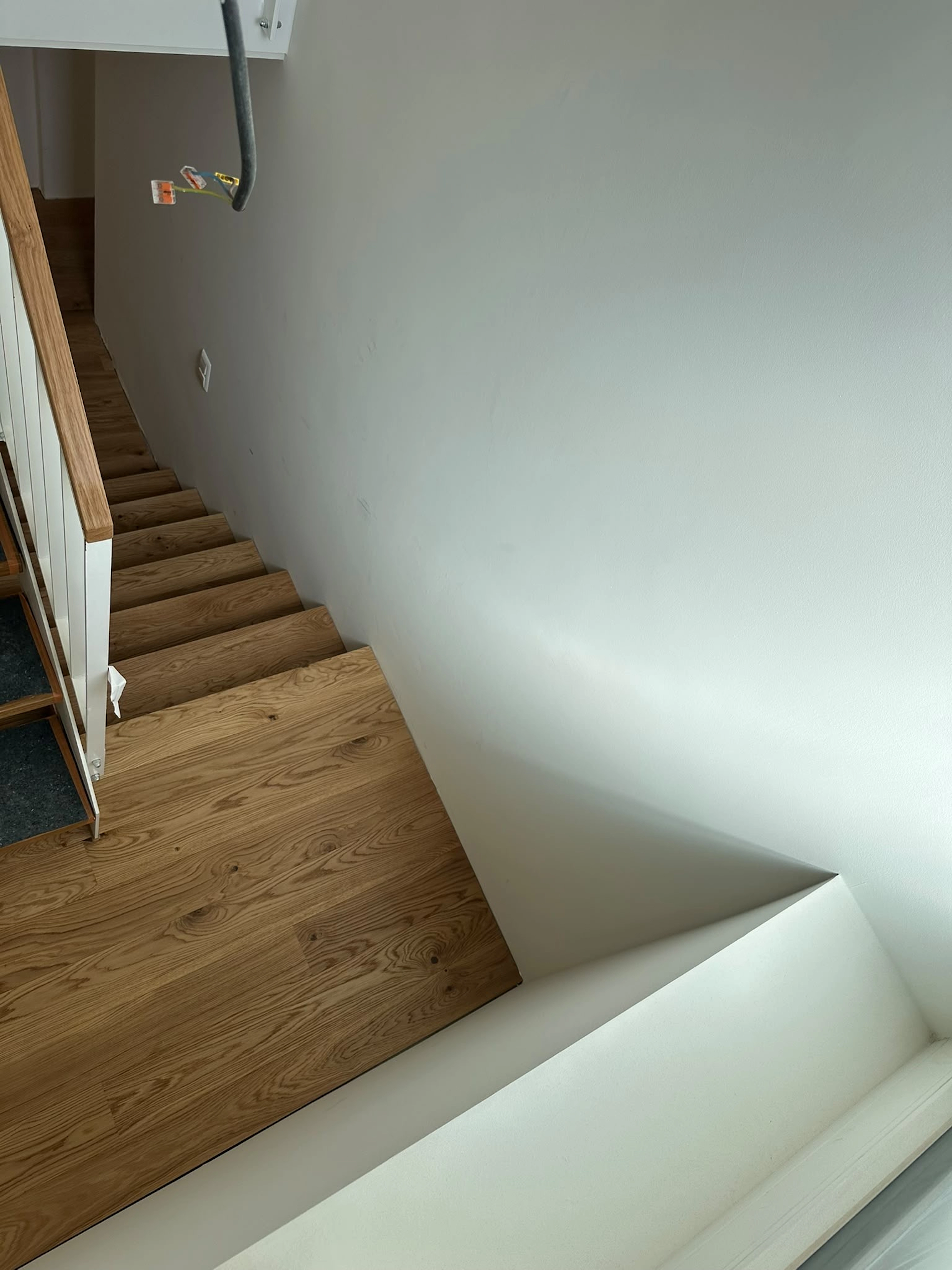 Perspektive vom Treppenpodest aus Parkettdielen auf mittlerer Ebene, mit Blick nach unten auf einen geraden Treppenlauf. Der Podestboden, gefertigt aus breiten Parkett Saussele-Landhausdielen, bildet den fließenden Materialübergang. Man sieht die unfertige Wand, einen freien Draht und den neuen Handlauf. Die Treppe nach unten hat standardmäßig separate Massivholz-Trittstufen, die den Unterschied zur maßgefertigten Parkettintegration betonen.