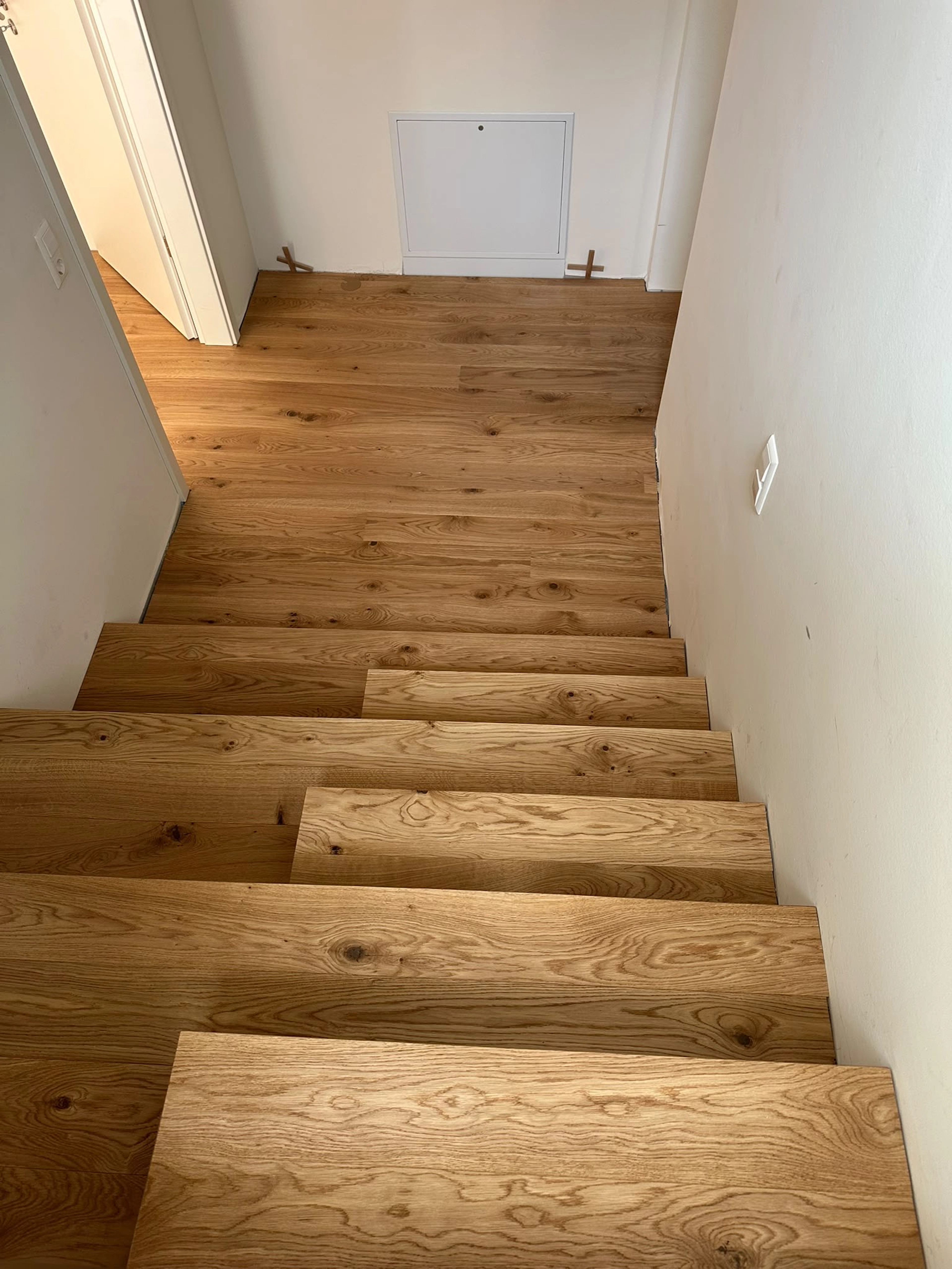 Weitwinkel-Aufnahme vom Treppenfuß, die den Beginn einer maßgefertigten Treppe zeigt. Die Stufen (Tritt- und Setzstufen) wurden direkt aus den breiten Parkett Saussele-Landhausdielen gefertigt und führen zu einem Podest mit demselben Parkettboden. Man sieht die unfertige, weiß gestrichene Wand und Floor-Spanner, die den fließenden Materialübergang betonen.