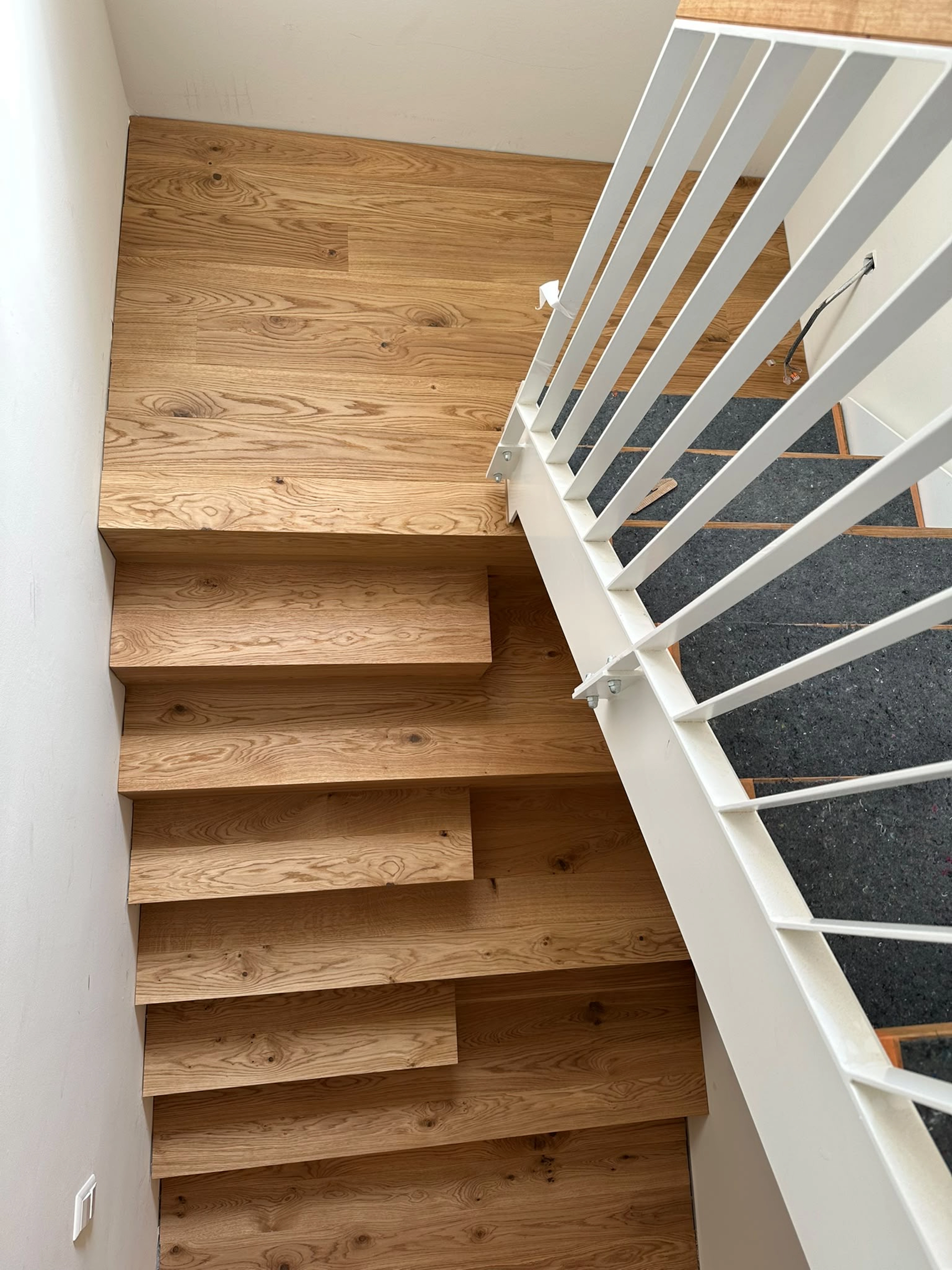 Flache Überkopf-Perspektive eines Treppenpodests auf mittlerer Ebene, das vollständig mit Parkett Saussele-Landhausdielen belegt ist. Eine moderne, weiß gestrichene Lamellen-Balustrade aus Metall begrenzt den Bereich. Das Podest bildet den Übergang zu einer zweiten Treppe mit weißem Metallrahmen, deren Stufen mit dunklen Schutzmatten bedeckt sind.