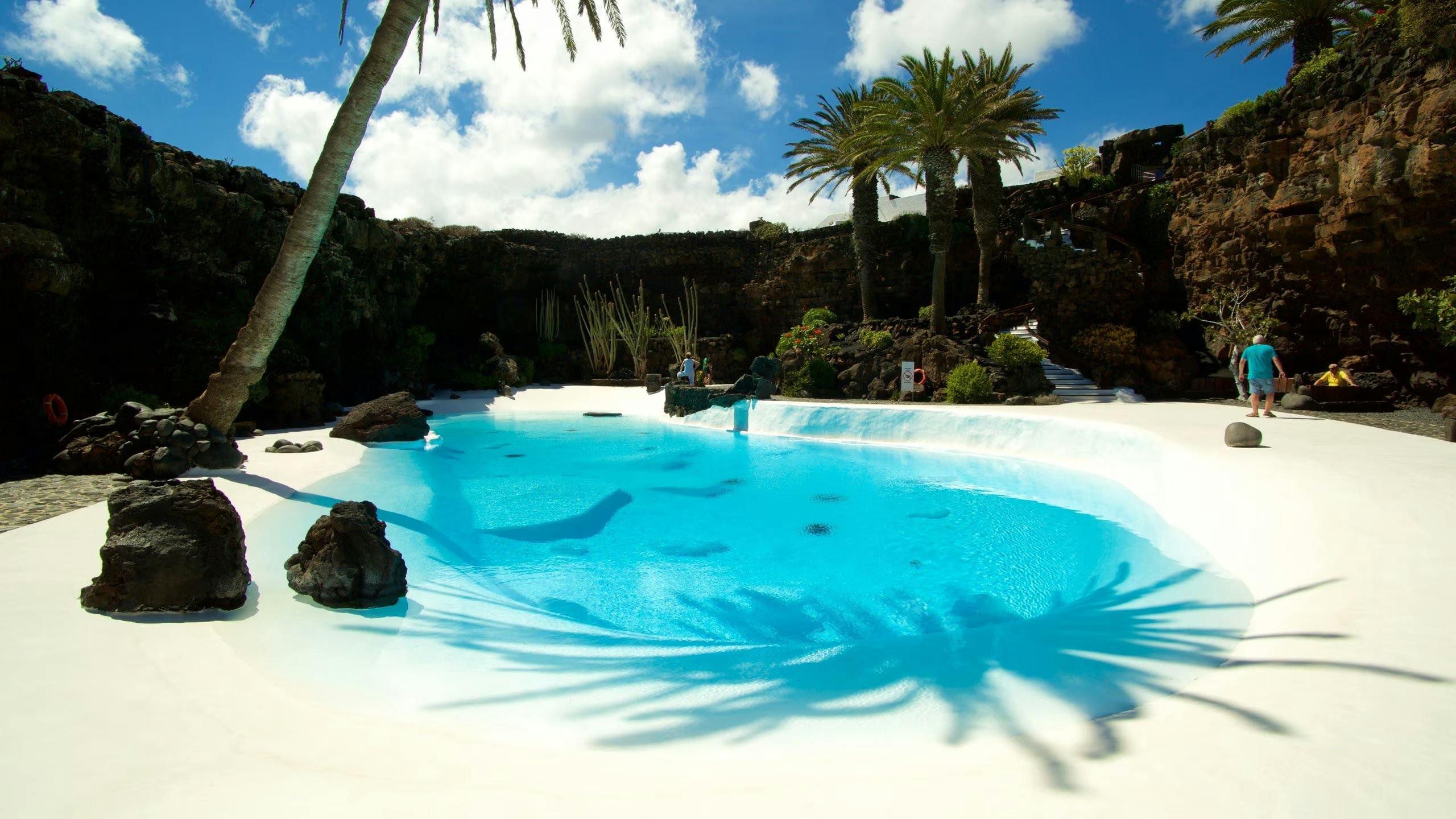 The chamber is illuminated by natural light filtering in through openings in the rock, creating a dramatic contrast between the dark volcanic stone and the bright blue of the pool.