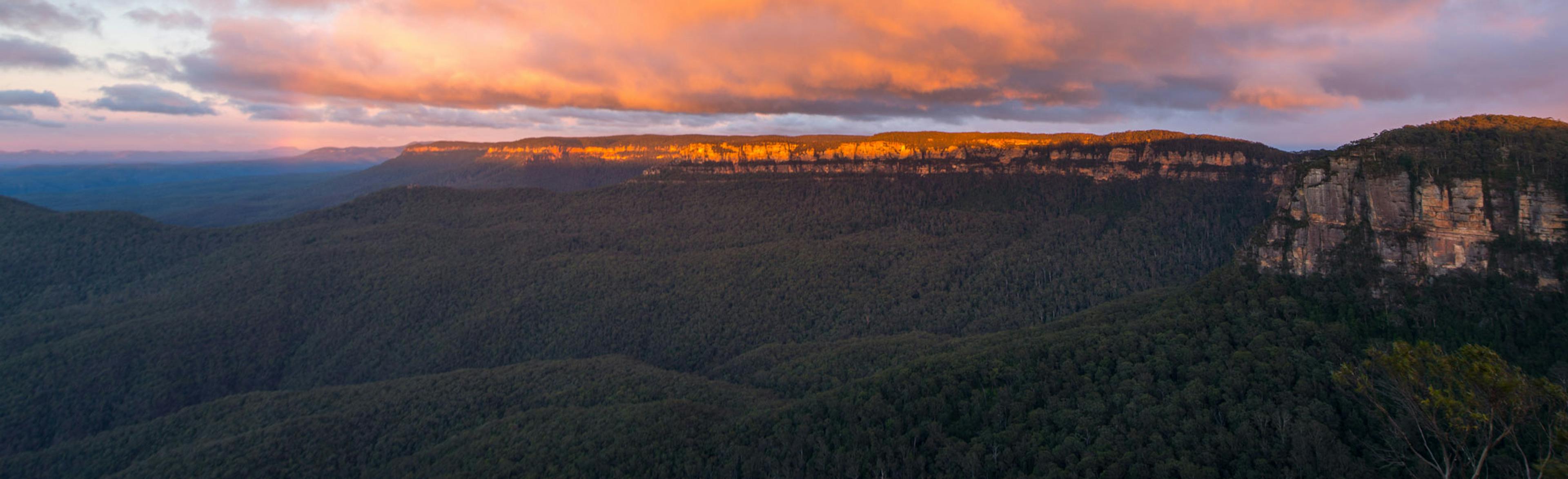 Panoramic view of a vast forested valley at sunset, with dramatic cliffs on the right. The sky is filled with colorful clouds, casting a warm orange glow over the landscape.