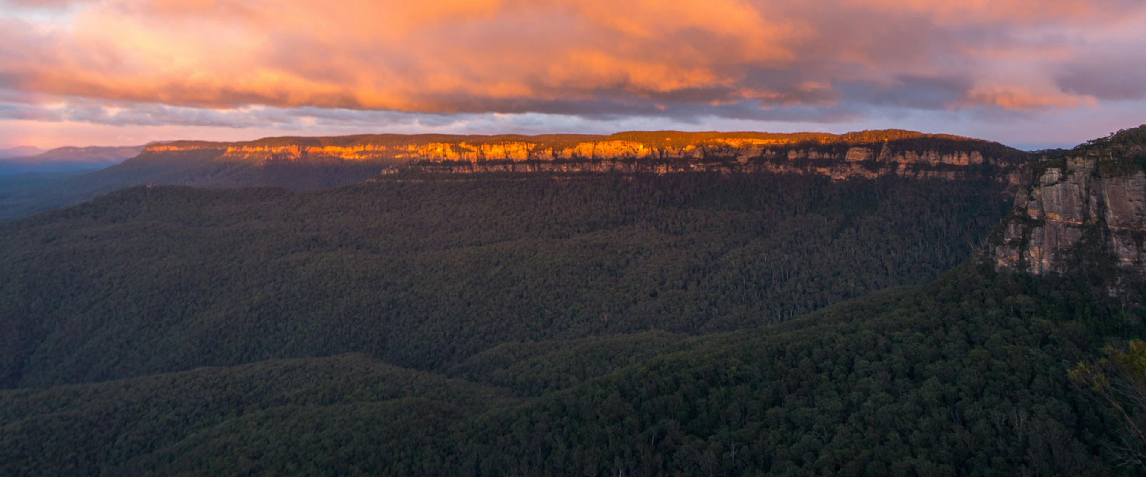 Panoramic view of a vast forested valley at sunset, with dramatic cliffs on the right. The sky is filled with colorful clouds, casting a warm orange glow over the landscape.