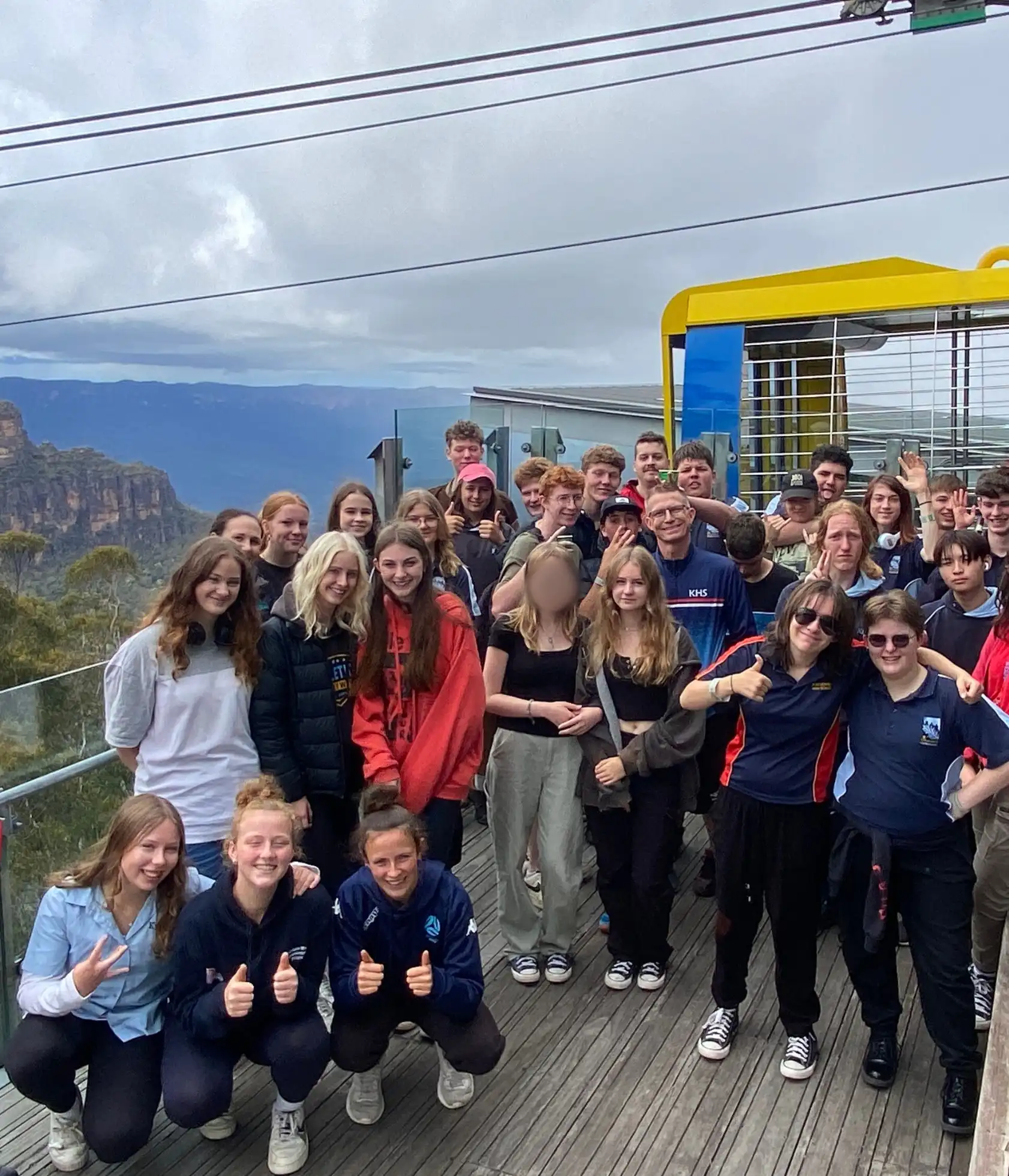 A group of people posing for a photo on an outdoor platform with mountainous scenery in the background. Some are wearing casual clothes while others are in uniforms. The atmosphere appears joyful and relaxed.