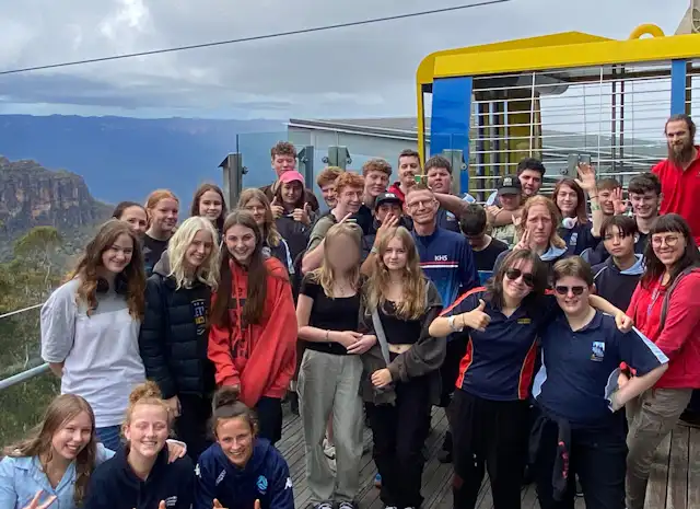 A group of people posing for a photo on an outdoor platform with mountainous scenery in the background. Some are wearing casual clothes while others are in uniforms. The atmosphere appears joyful and relaxed.