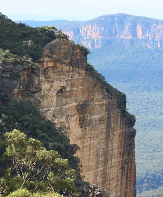 A steep rocky cliff face covered with sparse vegetation overlooks a vast valley of dense forest. In the background, more rugged cliffs rise under a partly cloudy sky, creating a striking and serene natural landscape.