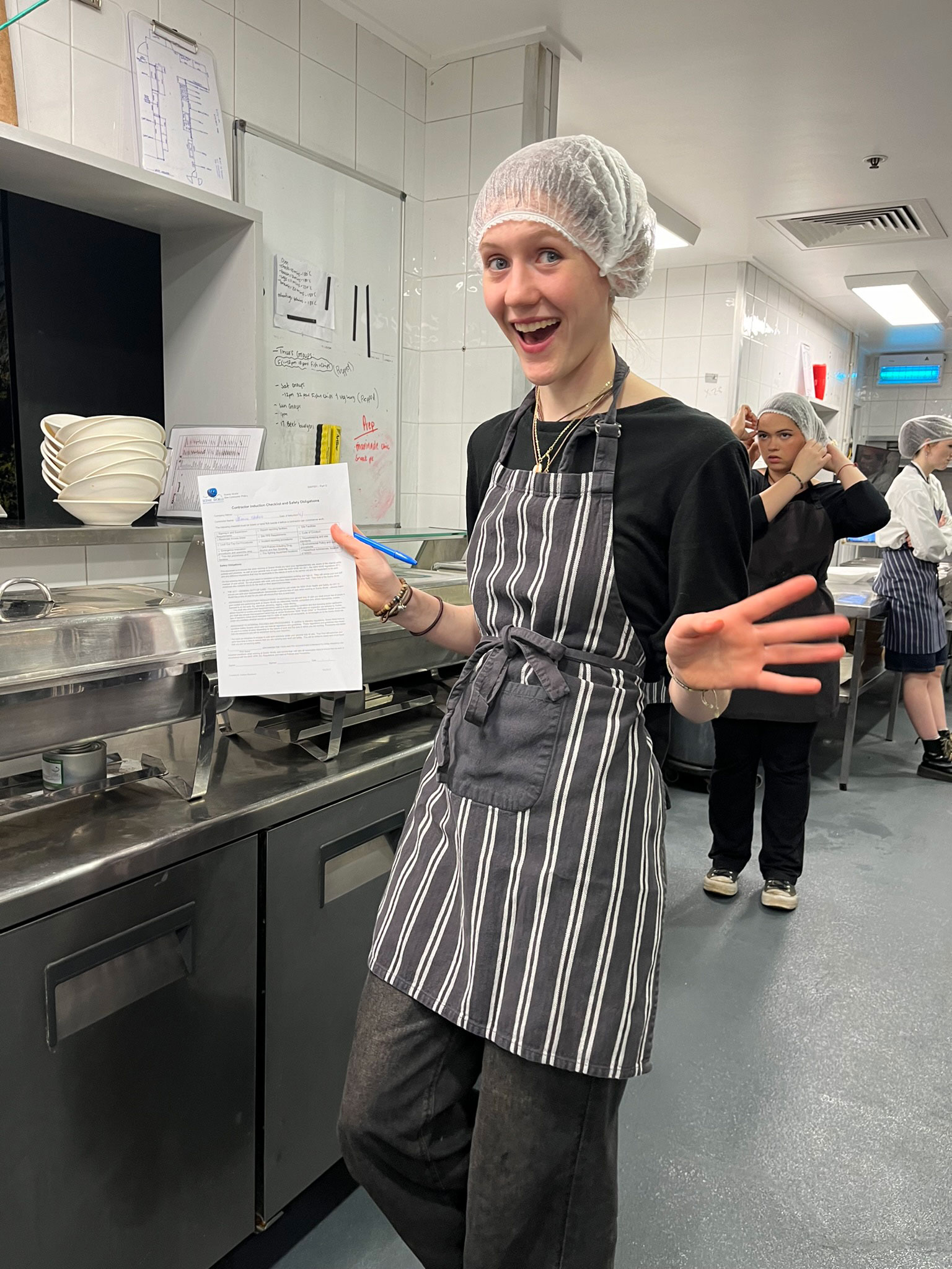 A person in a striped apron and hairnet smiles and holds a sheet of paper in a commercial kitchen. In the background, another person in similar attire is visible near the sink area. Whiteboards and kitchen equipment are also present.