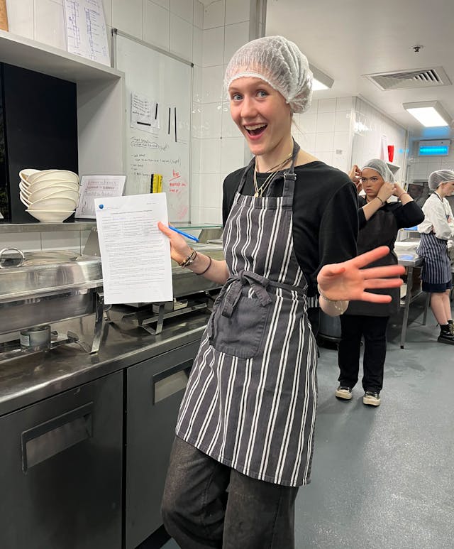 A person in a striped apron and hairnet smiles and holds a sheet of paper in a commercial kitchen. In the background, another person in similar attire is visible near the sink area. Whiteboards and kitchen equipment are also present.