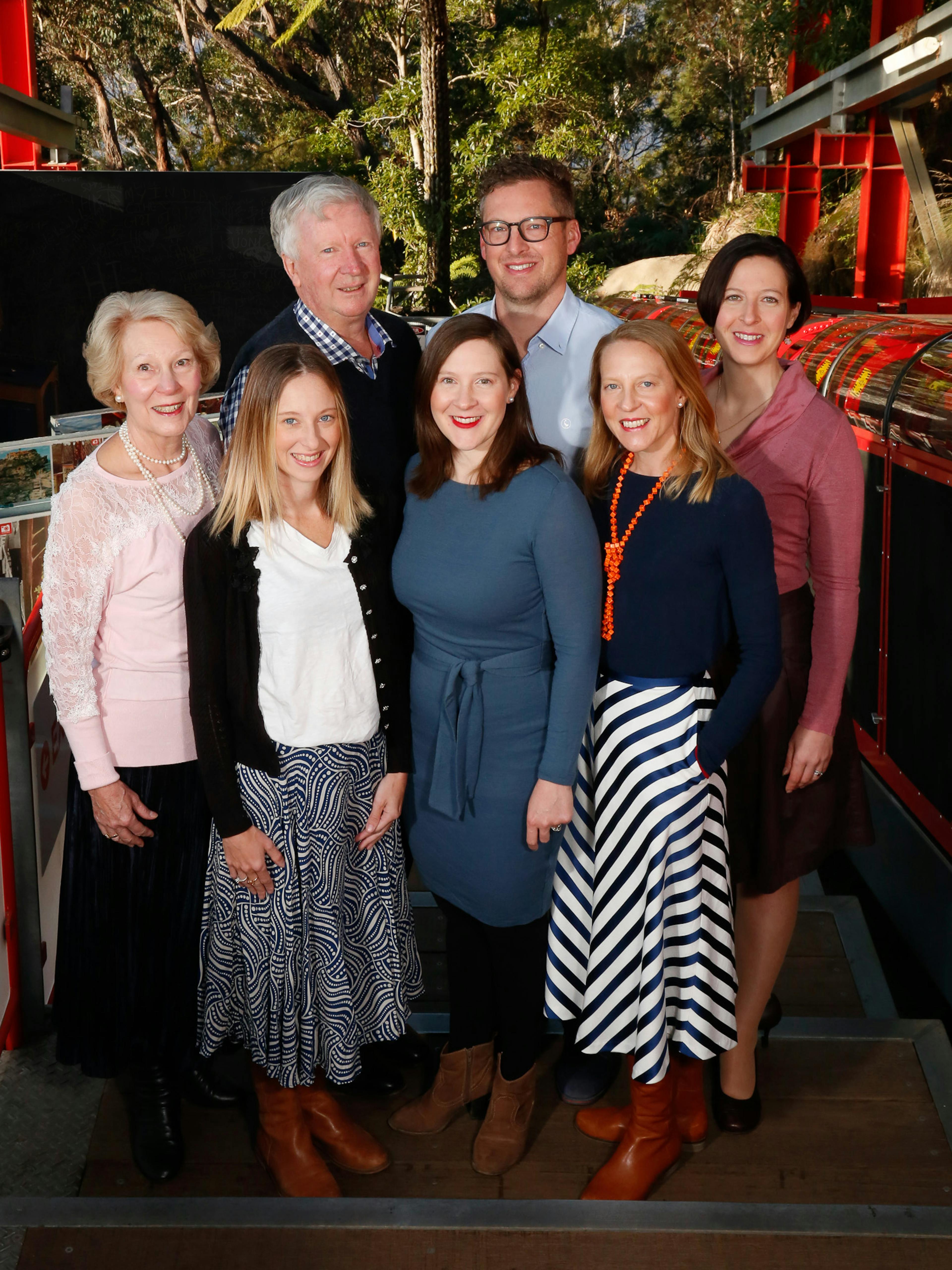 A group of seven people from the Hammon family stand together on the Railway platform. They are dressed in casual, colorful clothing. Trees and red railings are visible in the background, suggesting an outdoor setting.