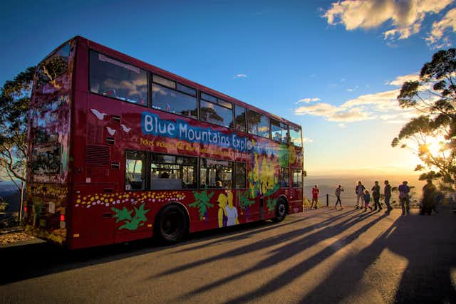 A red double-decker bus with a colorful design and Blue Mountains Explorer Bus written on the side is parked on a scenic lookout at sunset. People are standing nearby, taking in the view. Shadows from the trees and passengers stretch across the ground.