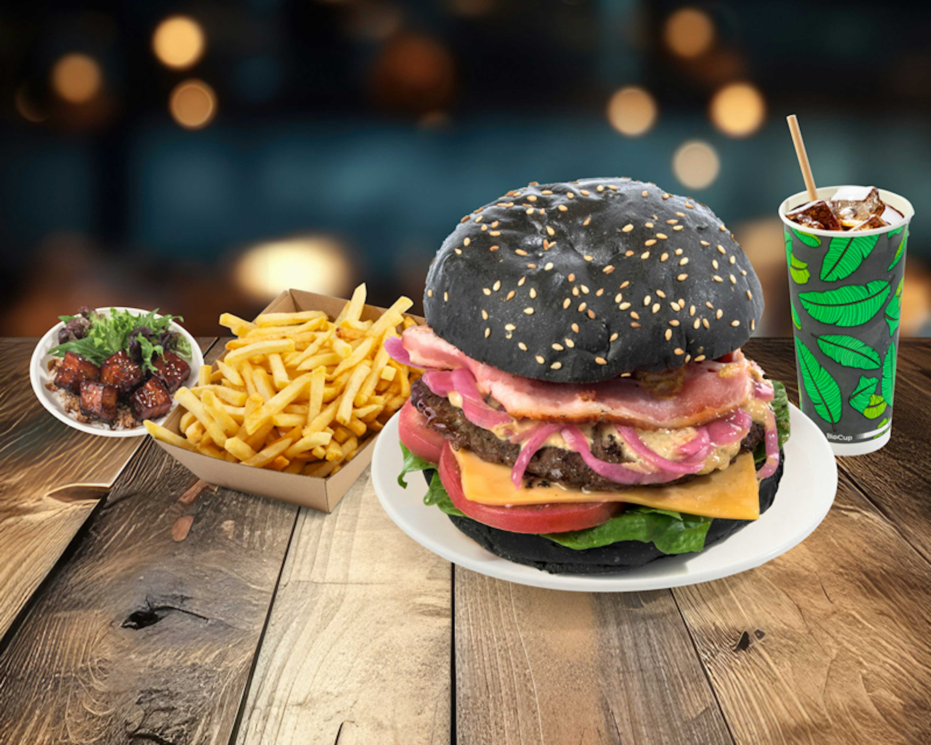 A burger with a black sesame seed bun, topped with bacon, cheese, and vegetables, is on a plate next to fries. Theres a salad with meat on the side and a cup with a drink in the background on a wooden table.