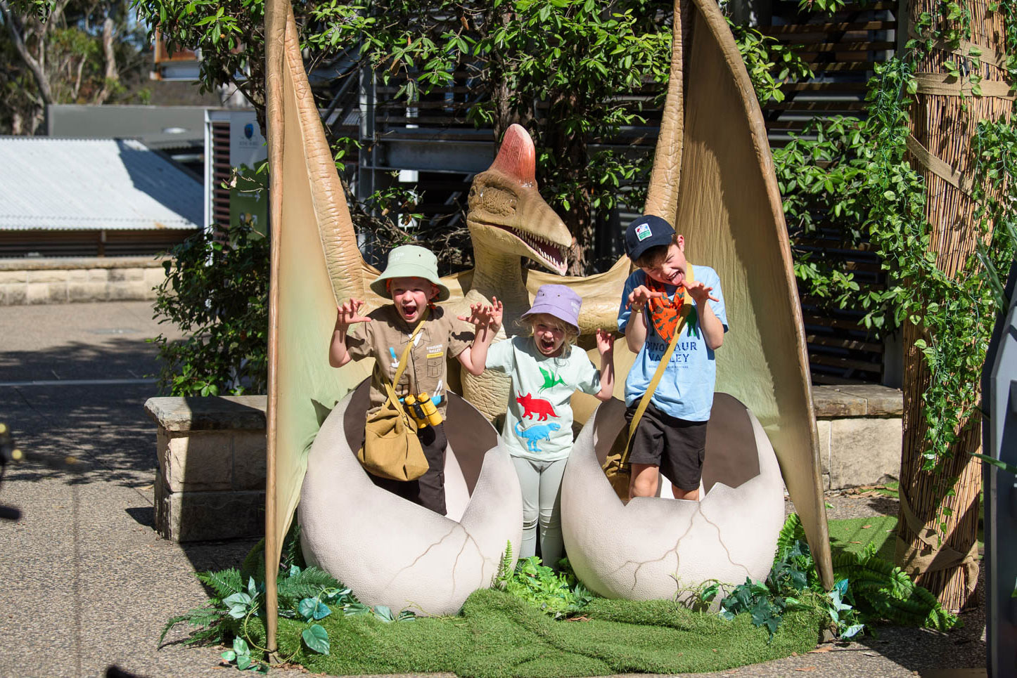 Three children, wearing hats and casual clothes, are posing playfully with pterosaur eggs at Scenic World. They are standing inside large fake eggs, surrounded by greenery. The pterosaur model has its wings spread wide behind them.