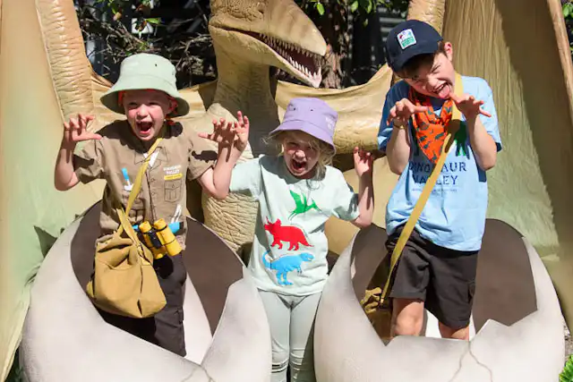 Three children, wearing hats and casual clothes, are posing playfully with pterosaur eggs at Scenic World. They are standing inside large fake eggs, surrounded by greenery. The pterosaur model has its wings spread wide behind them.