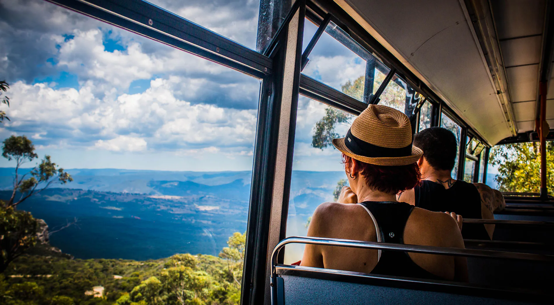 Two people sit inside a bus with large windows, enjoying a scenic view of lush green mountains under a partly cloudy sky. One person wears a straw hat while the other wears a cap, both gazing at the expansive landscape outside.