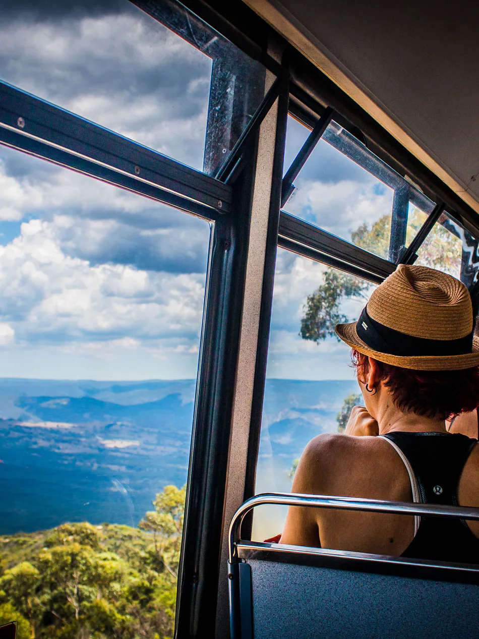 Two people sit inside a bus with large windows, enjoying a scenic view of lush green mountains under a partly cloudy sky. One person wears a straw hat while the other wears a cap, both gazing at the expansive landscape outside.