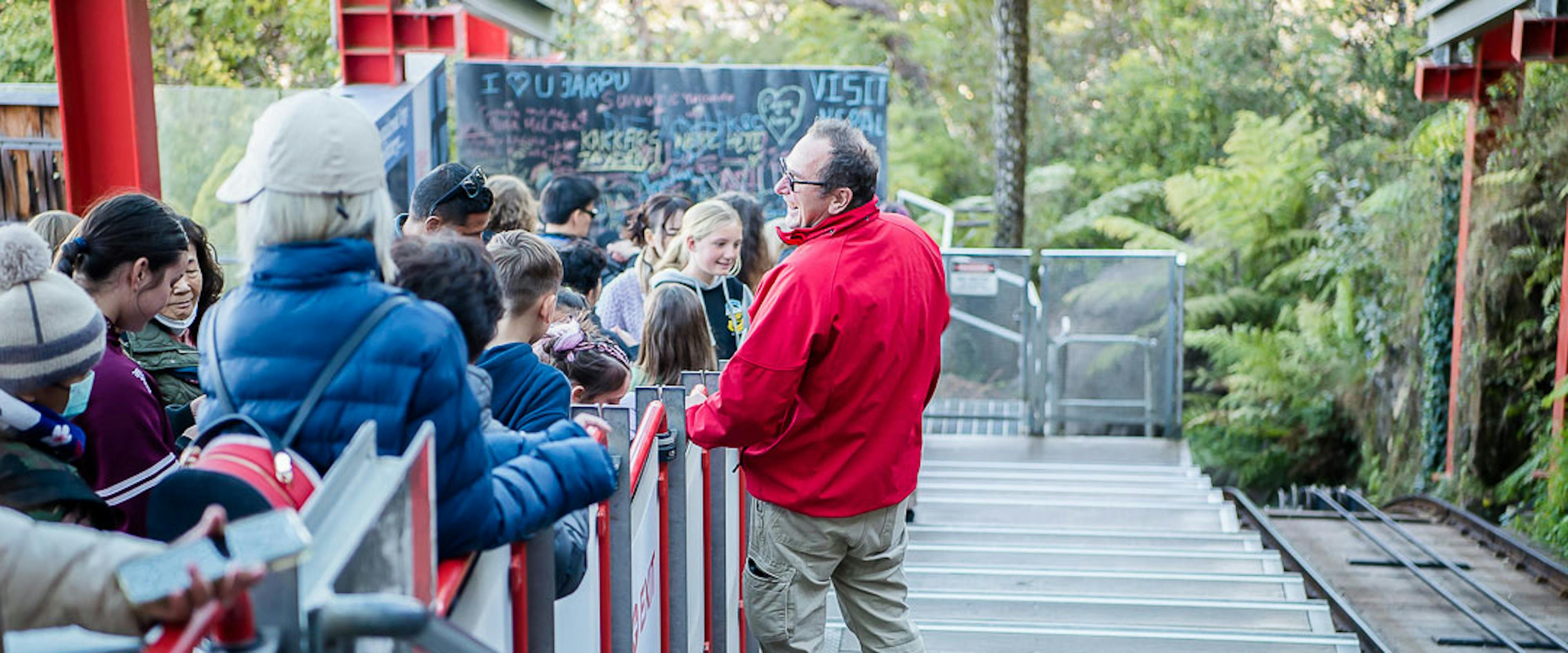 A group of people wait on a descending outdoor staircase. A man in a red jacket stands at the front, speaking to them. Lush greenery surrounds the area, and graffiti is visible on a wall in the background.