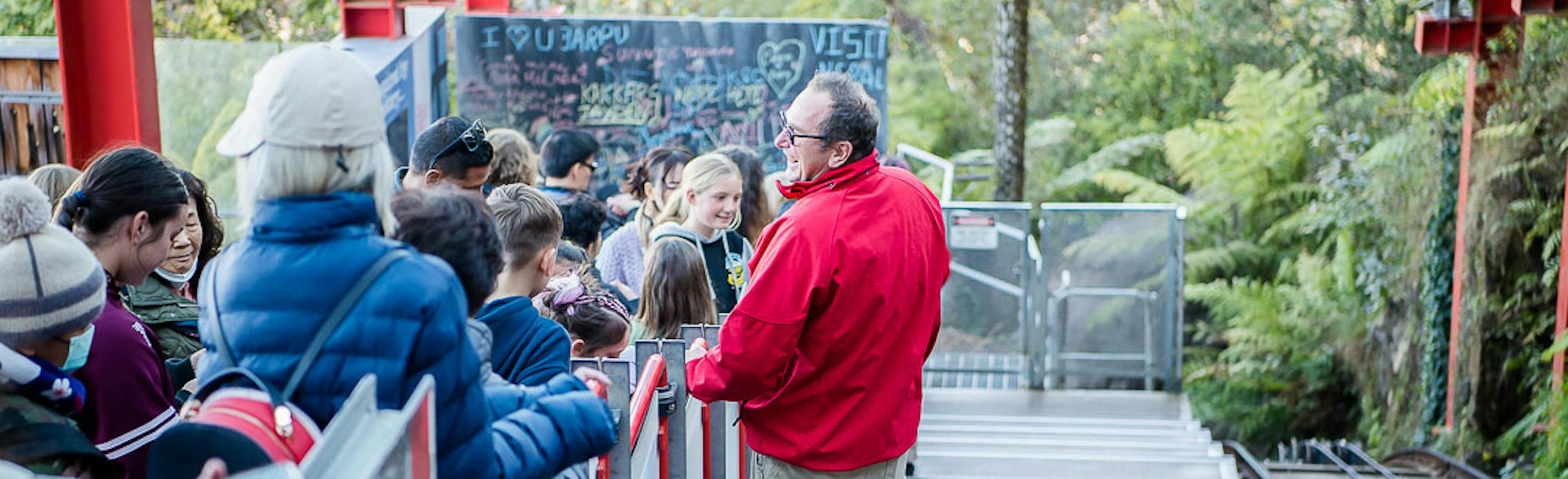 A group of people wait on a descending outdoor staircase. A man in a red jacket stands at the front, speaking to them. Lush greenery surrounds the area, and graffiti is visible on a wall in the background.