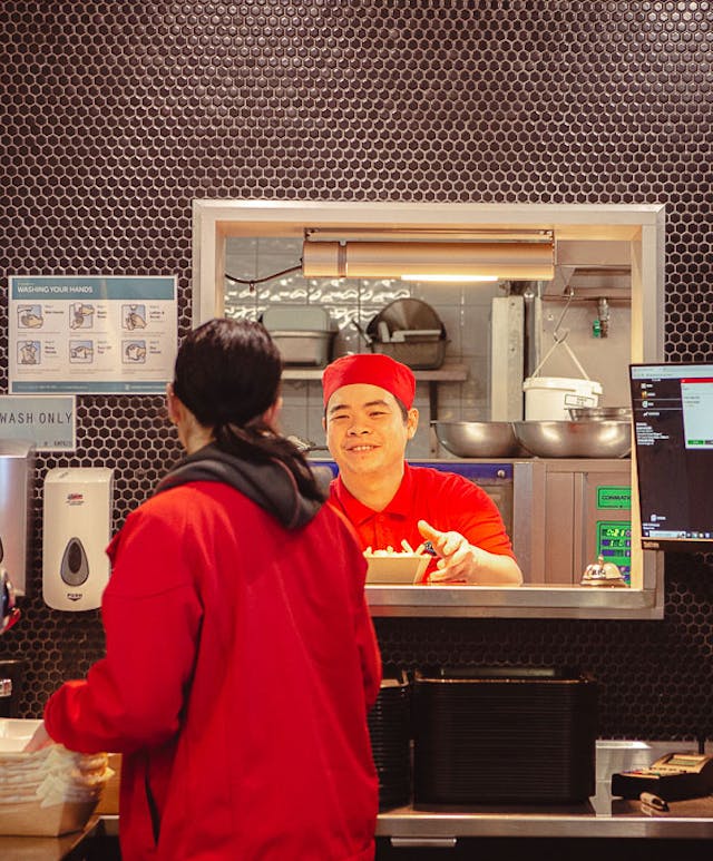 A person in a red uniform and cap stands behind a counter with a serving window, smiling at a customer. The setting is a modern food service area with various equipment and a drink dispenser visible.