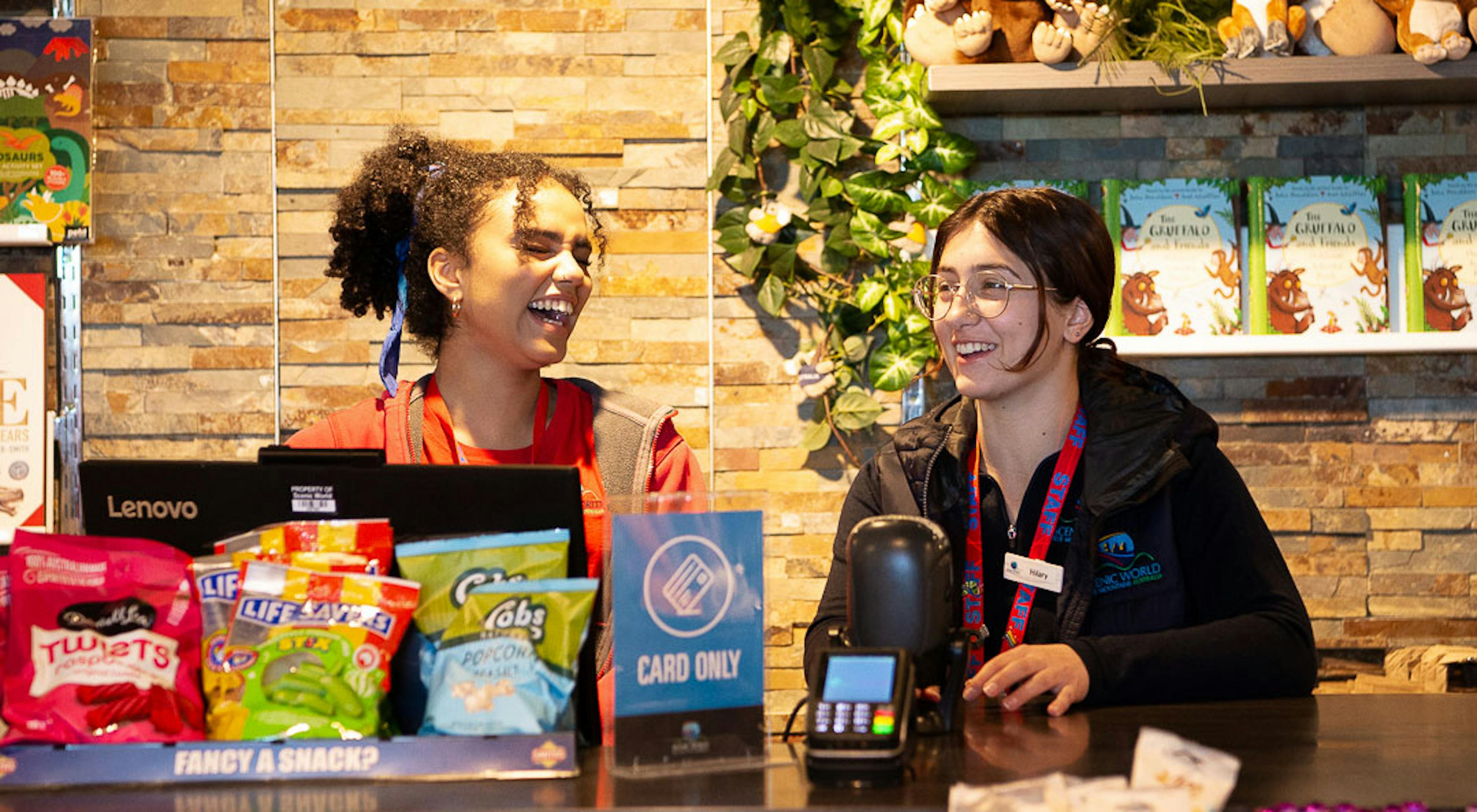 Two employees stand behind a counter in a store. They are smiling and chatting. In front of them are various snacks, and a sign that says Card Only. The background includes shelves with plush toys and green plants.