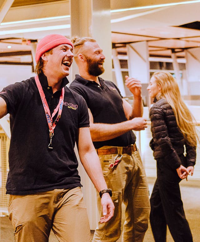 Three people wearing casual uniforms stand in a modern, indoor setting. Two of them are laughing, and one is wearing a red beanie. The area features metal railings and bright overhead lighting.