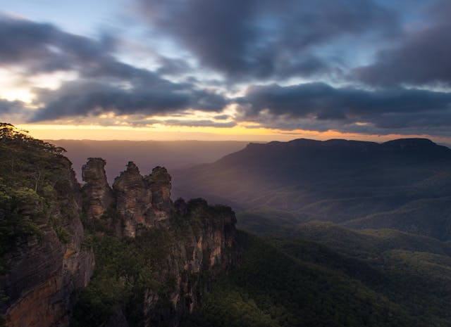 A dramatic sunrise over the Blue Mountains in Australia, featuring the iconic rock formation known as the Three Sisters. Dense green forests blanket the valleys below, under a sky filled with dark, rolling clouds.