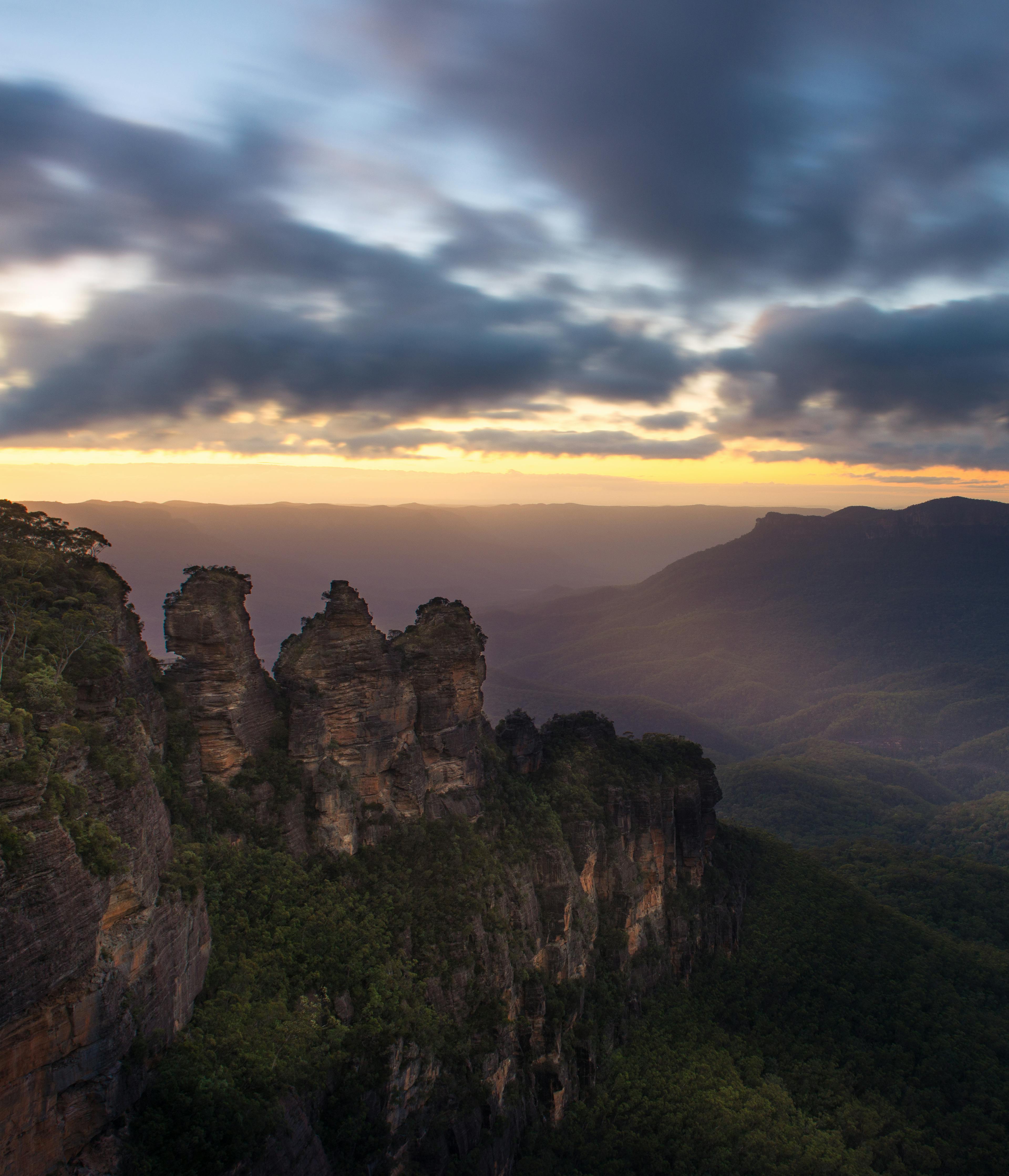 A dramatic sunrise over the Blue Mountains in Australia, featuring the iconic rock formation known as the Three Sisters. Dense green forests blanket the valleys below, under a sky filled with dark, rolling clouds.