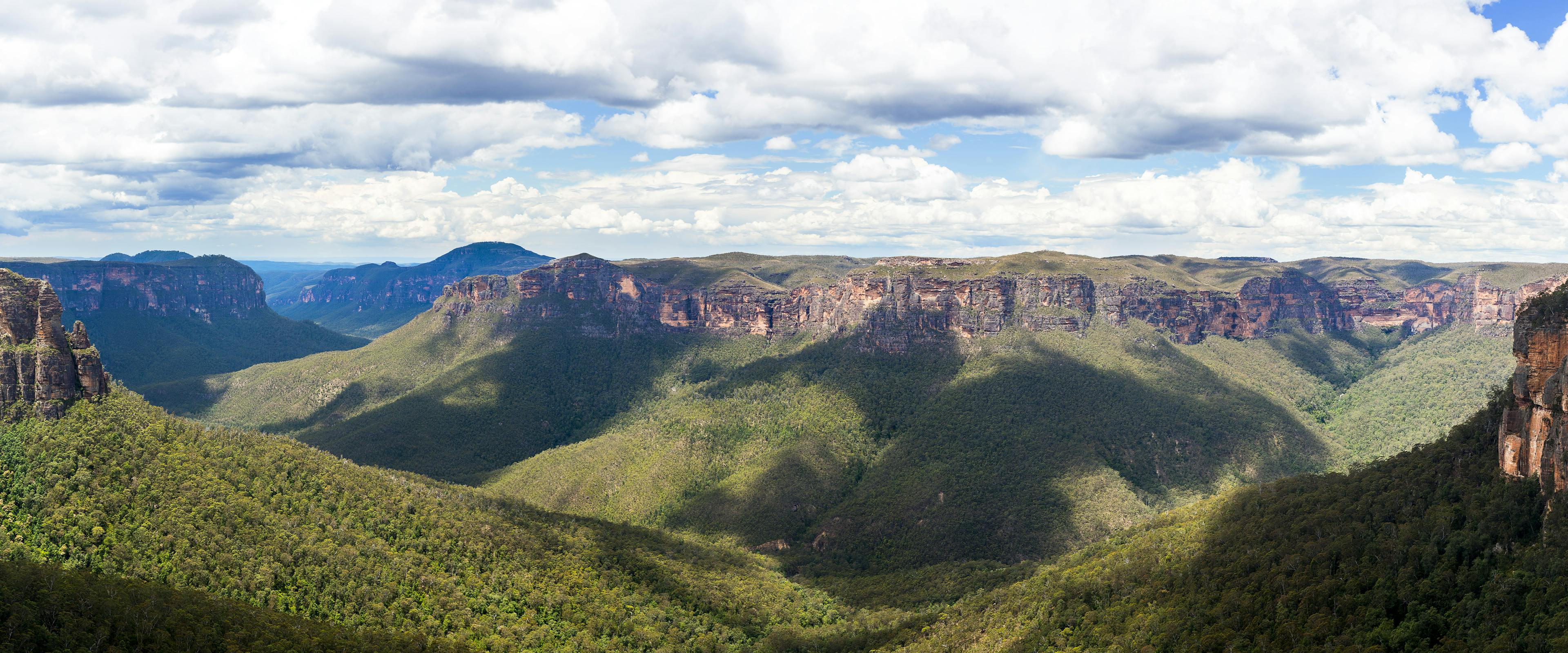 Panoramic view of a lush, green canyon with steep cliffs under a partly cloudy sky. Shadows from clouds create contrasting patterns on the forested landscape below.