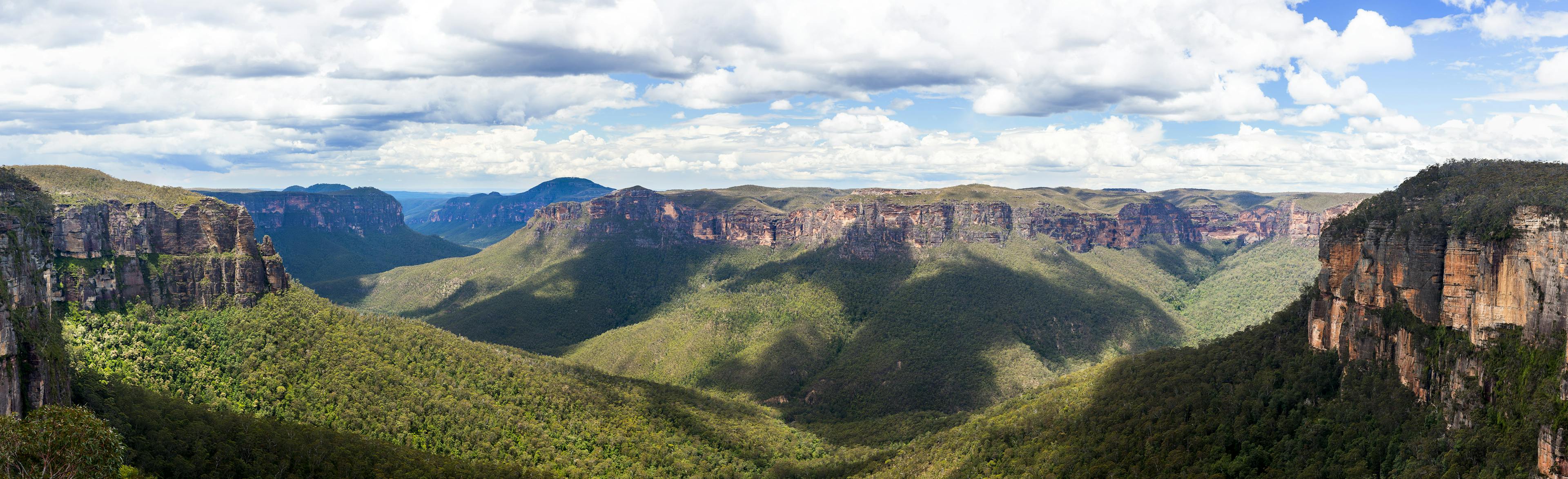 Panoramic view of a lush, green canyon with steep cliffs under a partly cloudy sky. Shadows from clouds create contrasting patterns on the forested landscape below.